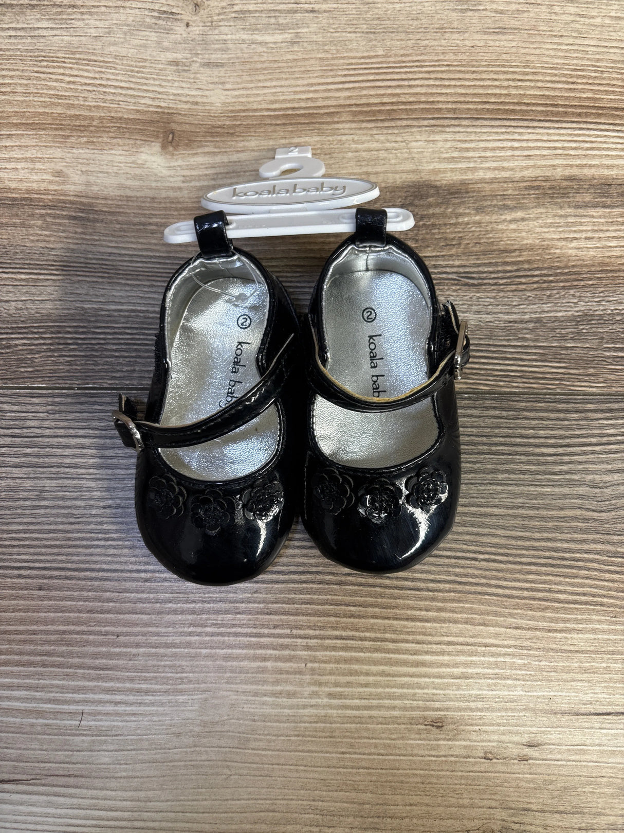 Top view of Koala Baby black patent Mary Jane flats size 2C with floral toe detail and buckle straps on wood background