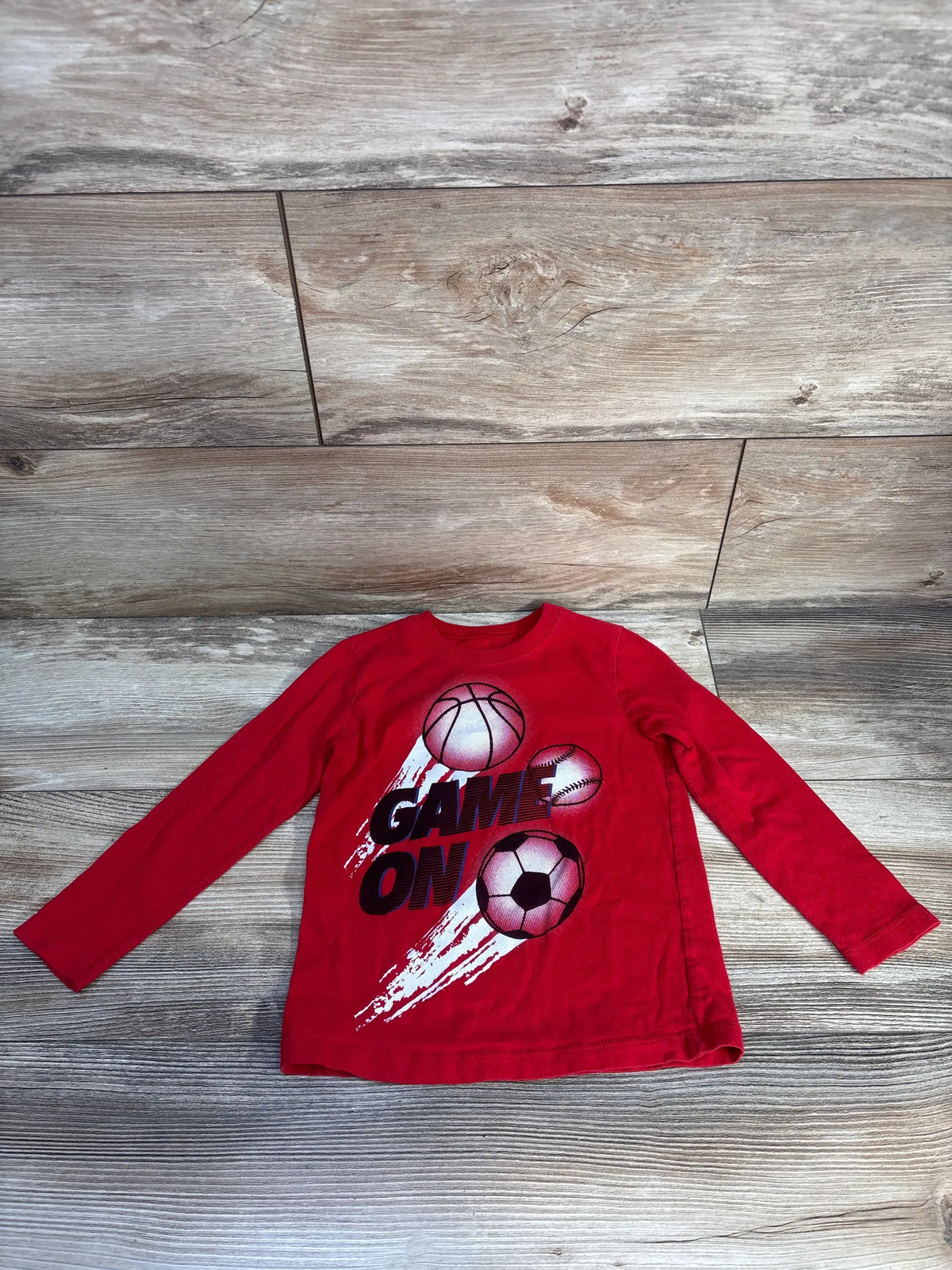 Front view of Carter’s toddler shirt showcasing red long sleeves and Game On sports ball graphic with wood grain background