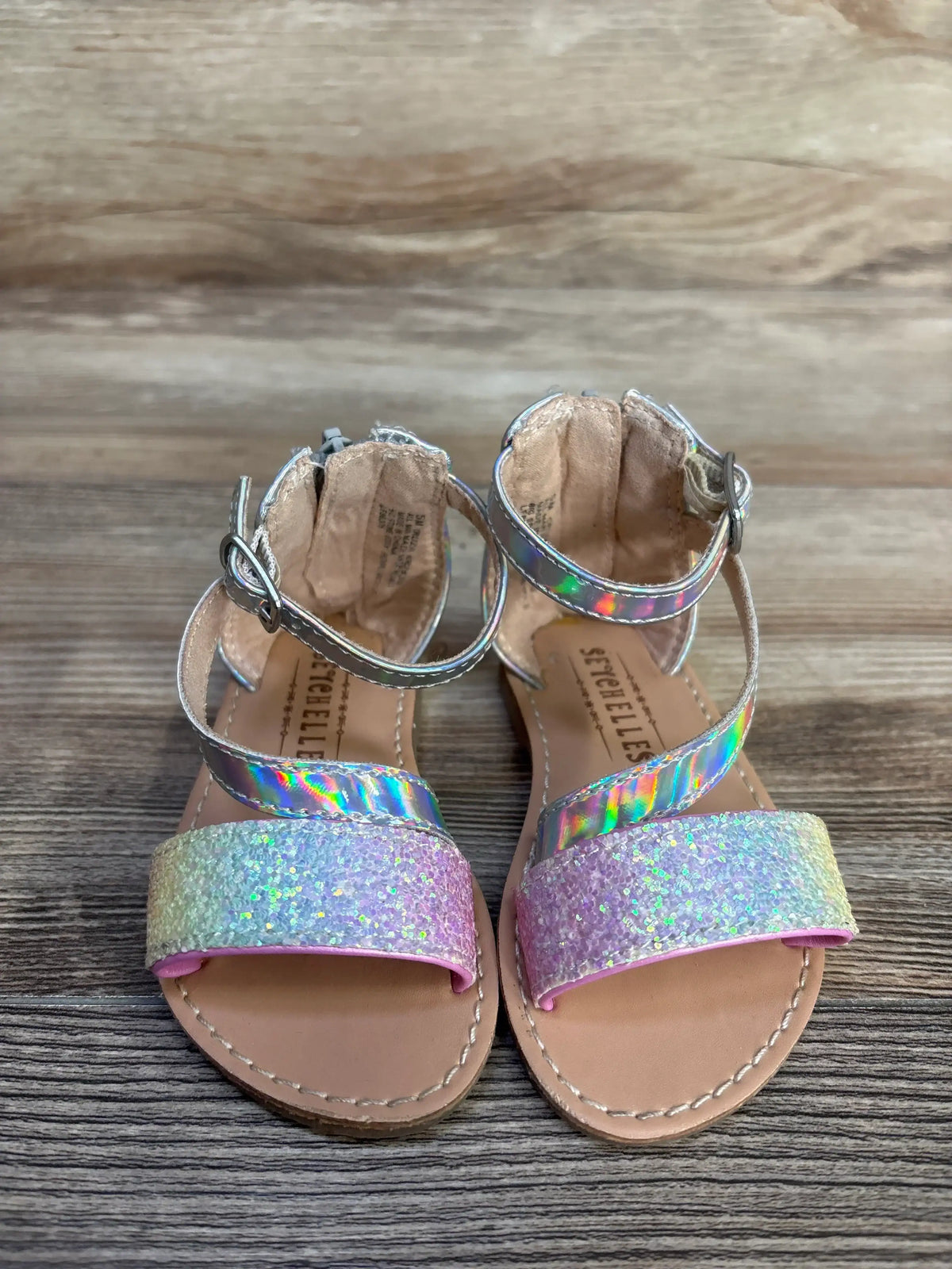 Top-down view of Seychelles toddler metallic sandals showing holographic ankle straps and pink iridescent glitter toe straps on wooden floor