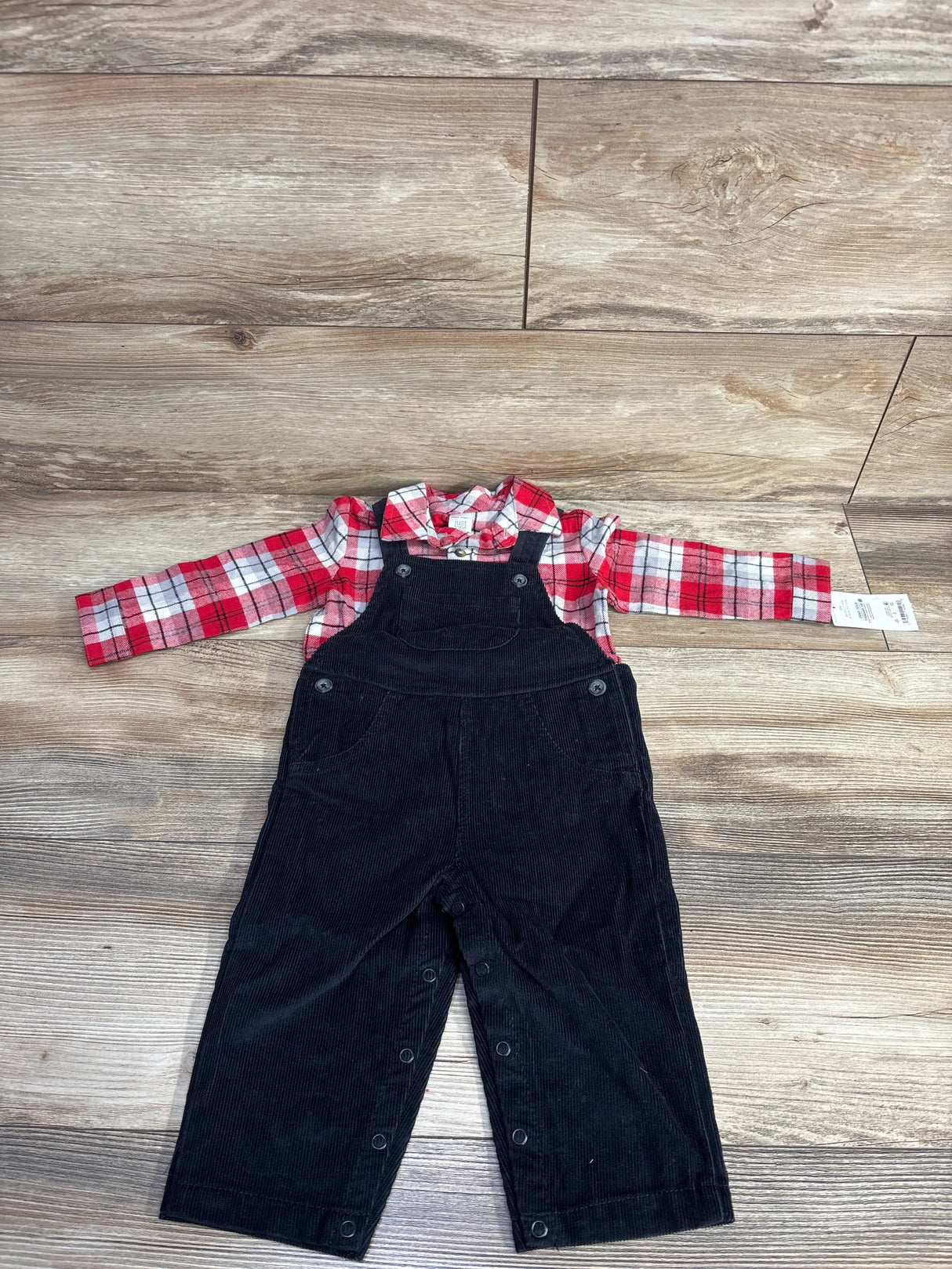 Baby outfit with red plaid shirt and black corduroy overalls on wood floor