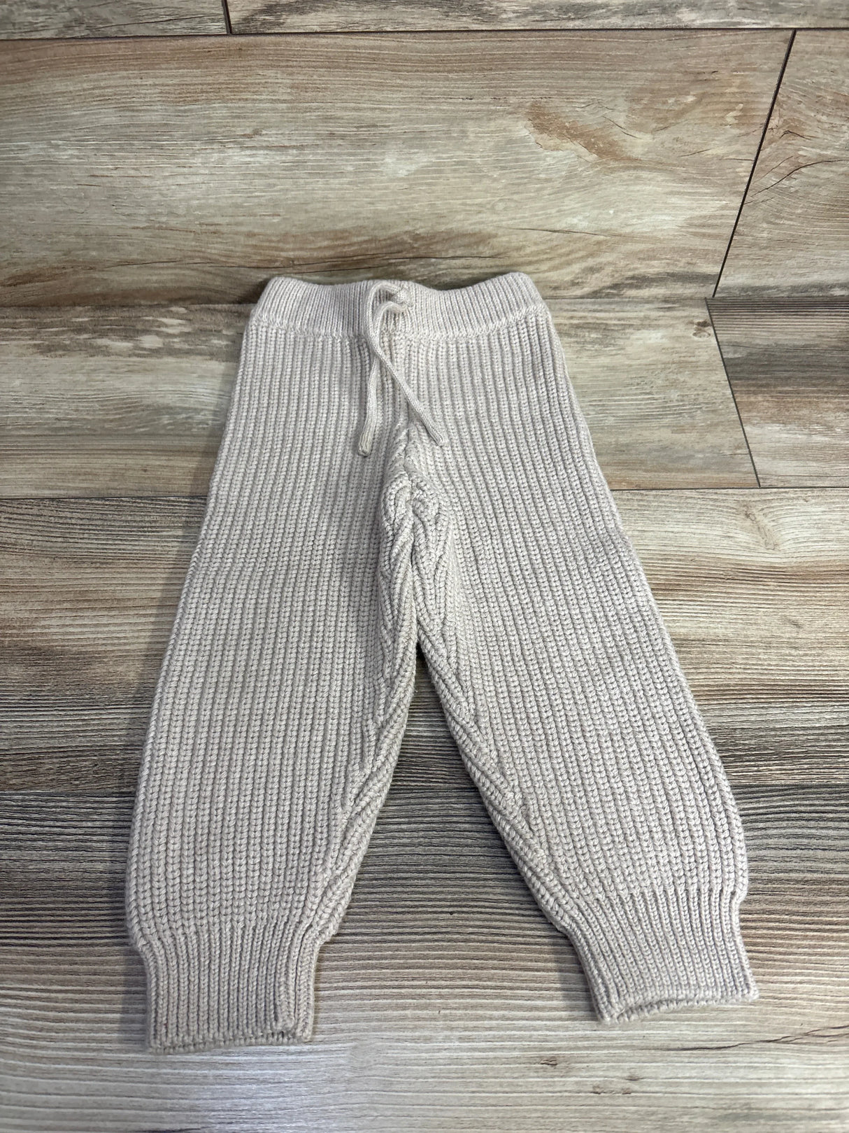 Beige knit toddler pants with drawstring waist and ribbed texture on wood background