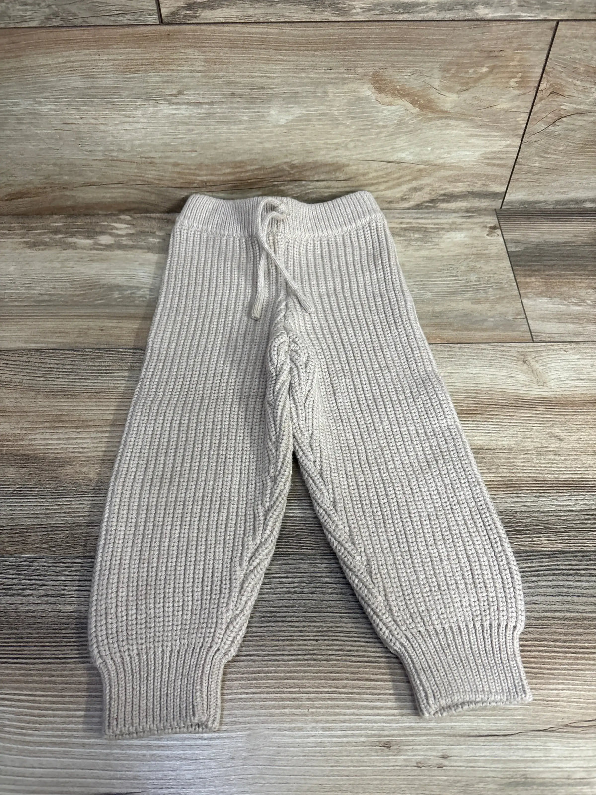 Beige knit toddler pants with drawstring waist and ribbed texture on wood background