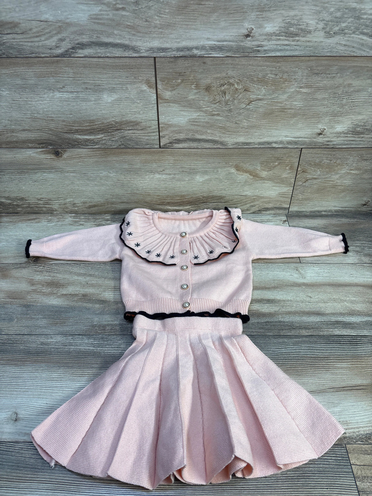Flat lay of pink knit button-front cardigan with ruffled embroidered collar and matching pleated skirt on wood floor