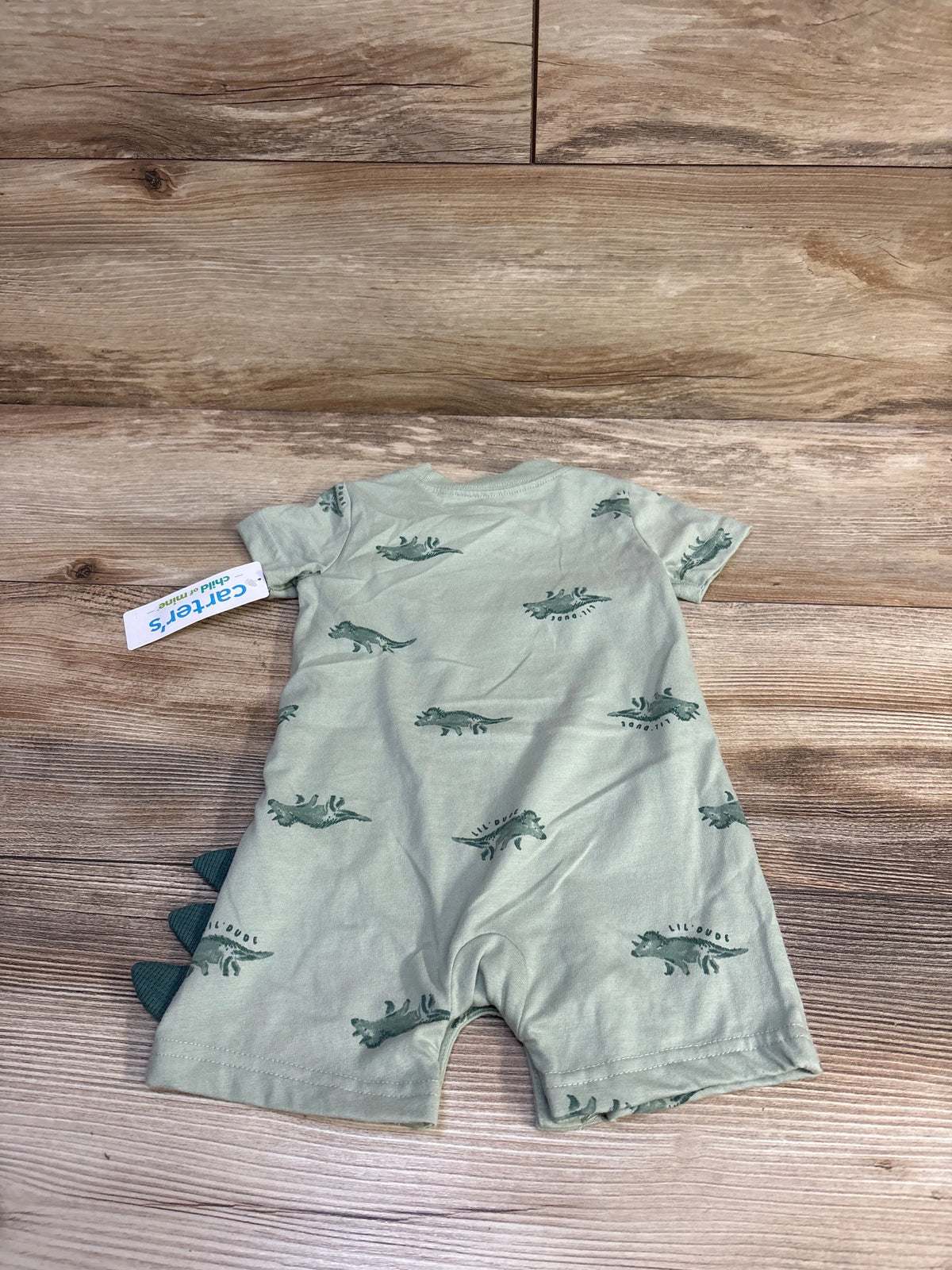 Back view of Child of Mine shortie romper in sage green with small dinosaur print and fabric spikes along left side on wood background
