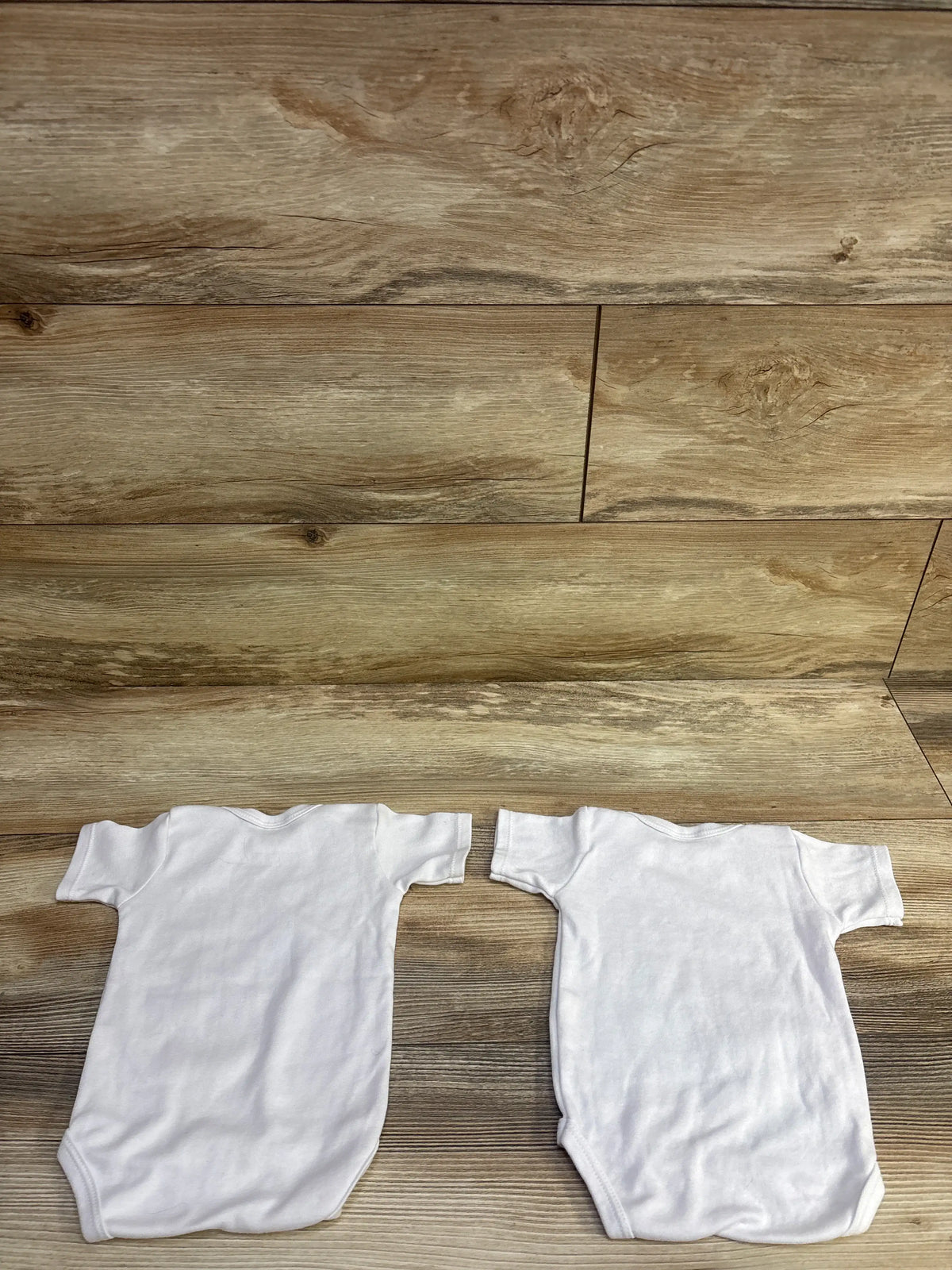 Top view of two white short-sleeve baby bodysuits showing rounded necklines on a wood-grain background
