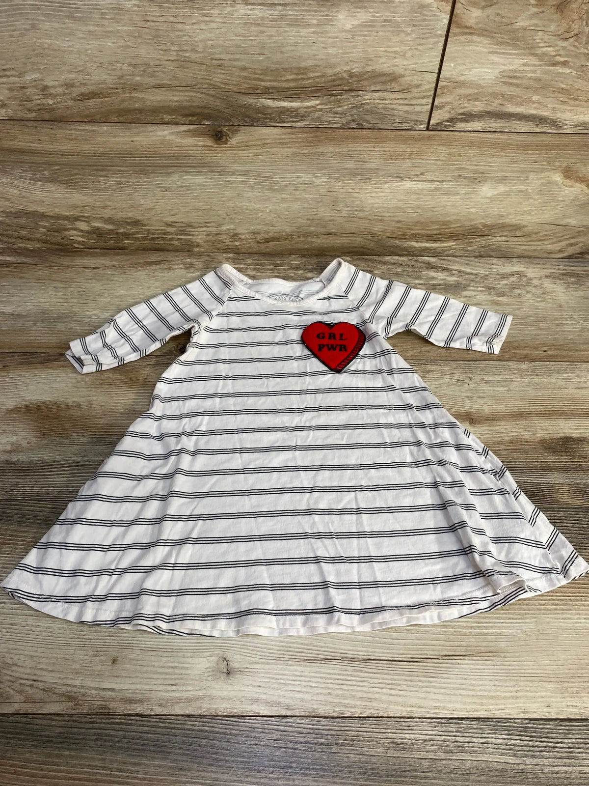 Flat lay of white and black striped toddler dress showcasing red heart patch reading GRL PWR on chest with wood plank background