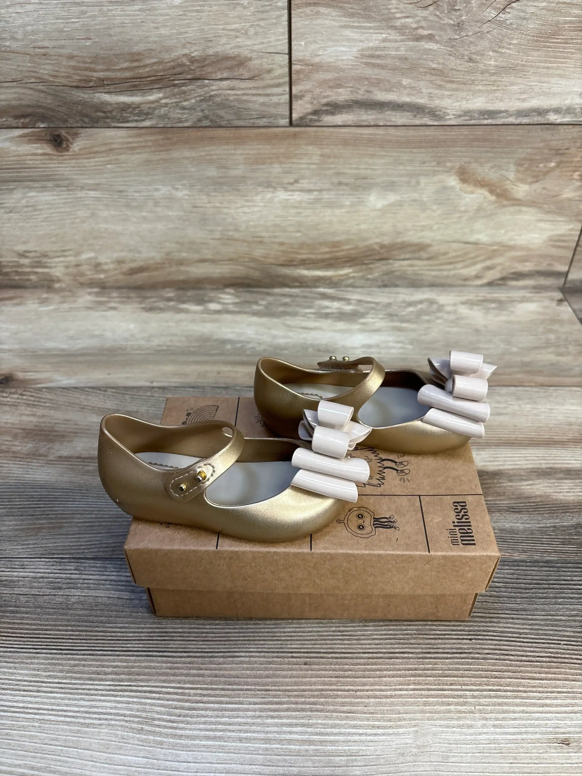 Gold toddler Mary Jane shoes with white bows on a cardboard box, wooden background