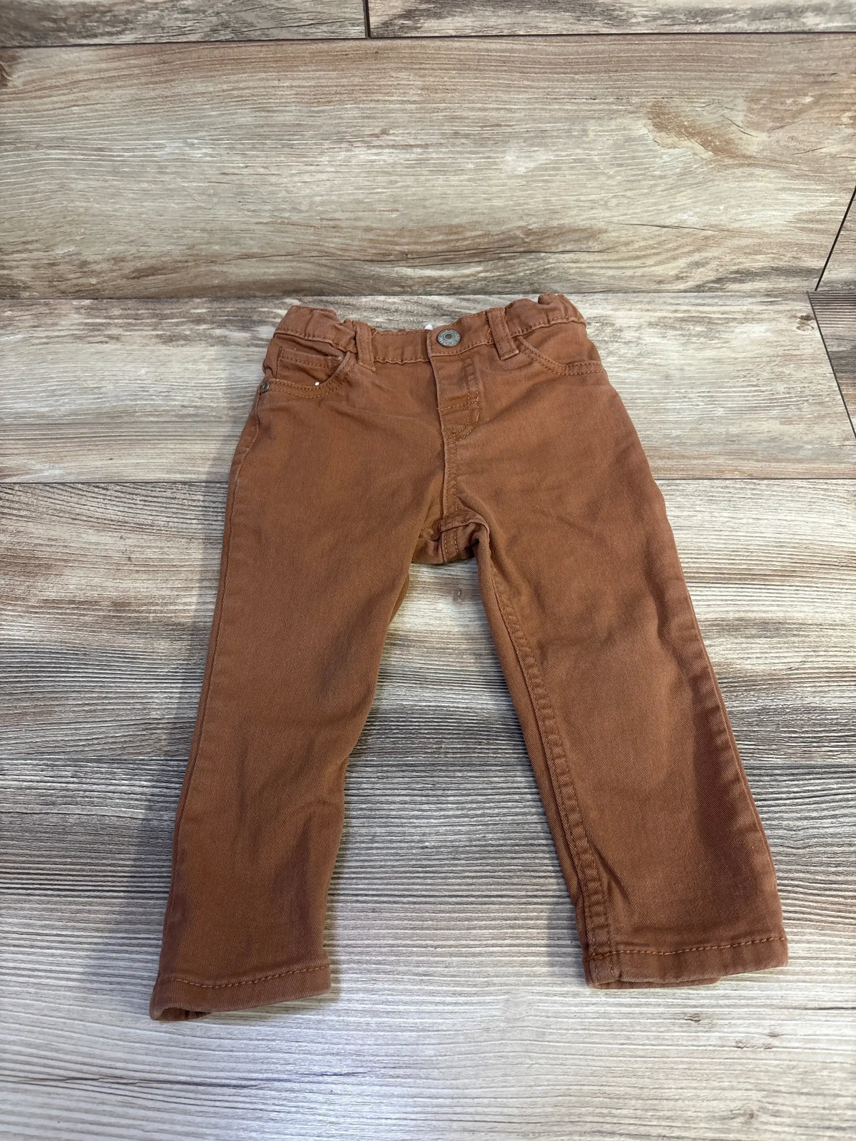 Front flat-lay of brown H&M twill baby pants (12m) showing elastic waist, front pockets, tapered legs on wood background