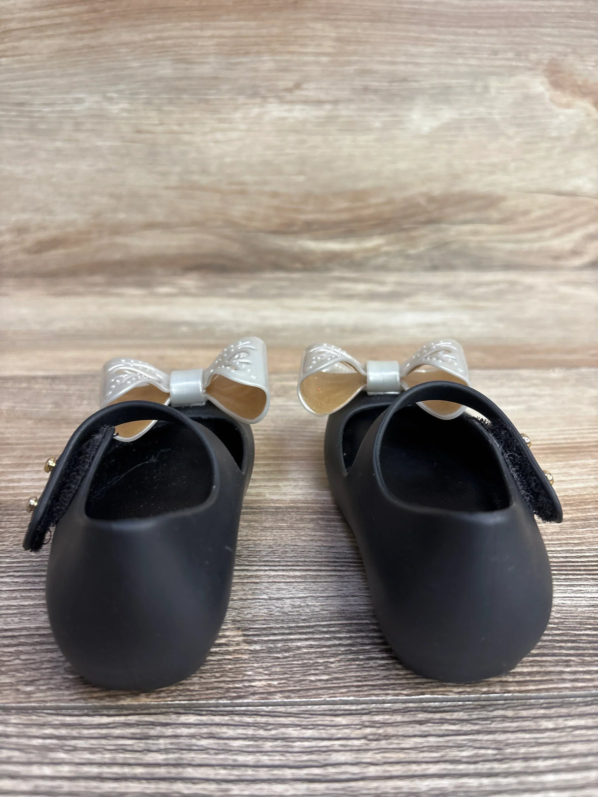 Front view of Mini Melissa Ultragirl Bow jelly flats in black size 5C showing silver bows and Mary Jane straps on wood background