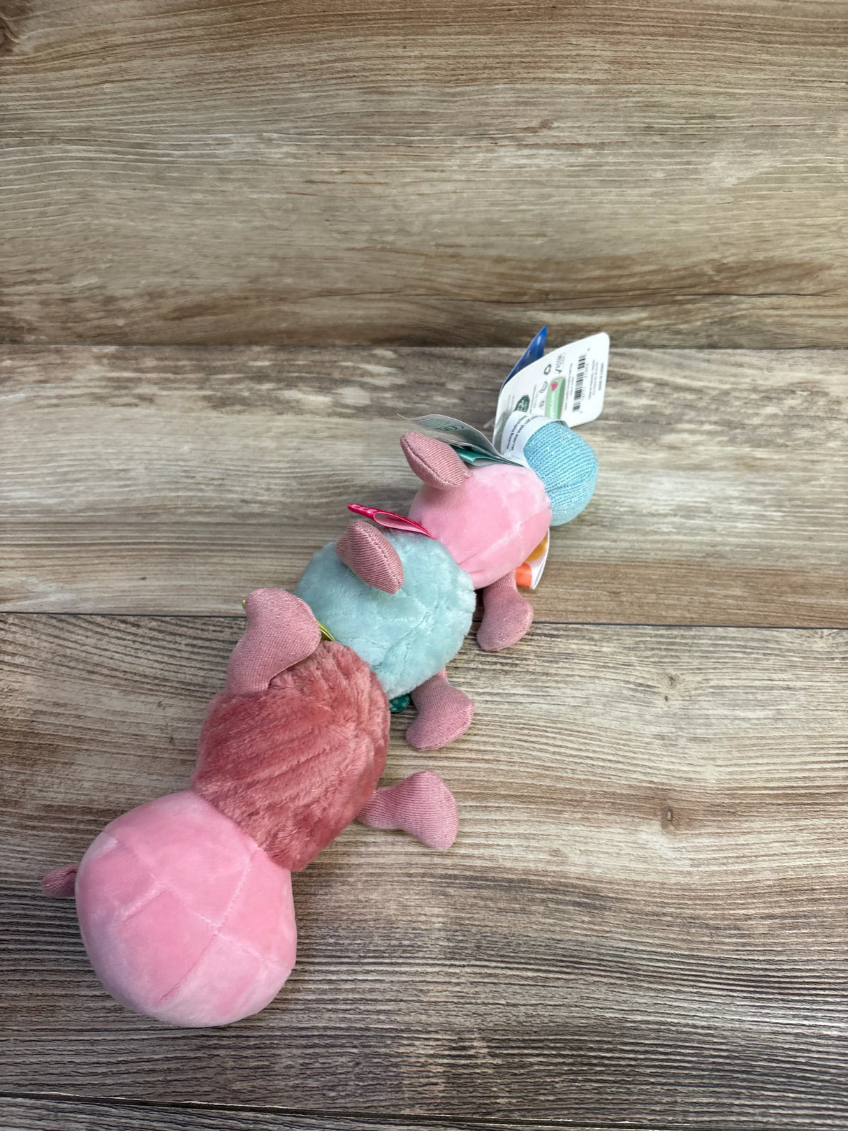 Angled view of Taggies Camilla Caterpillar toy showing pink and teal plush sections and colorful fabric tags on wood floor