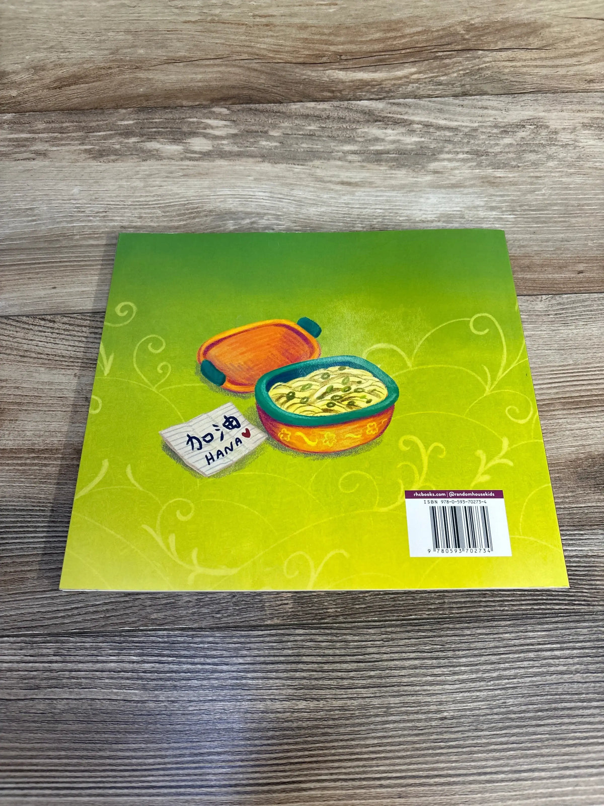 Flat lay of How We Say I Love You paperback showing green back cover with bento box illustration, barcode, and note ‘HANA’ on wood