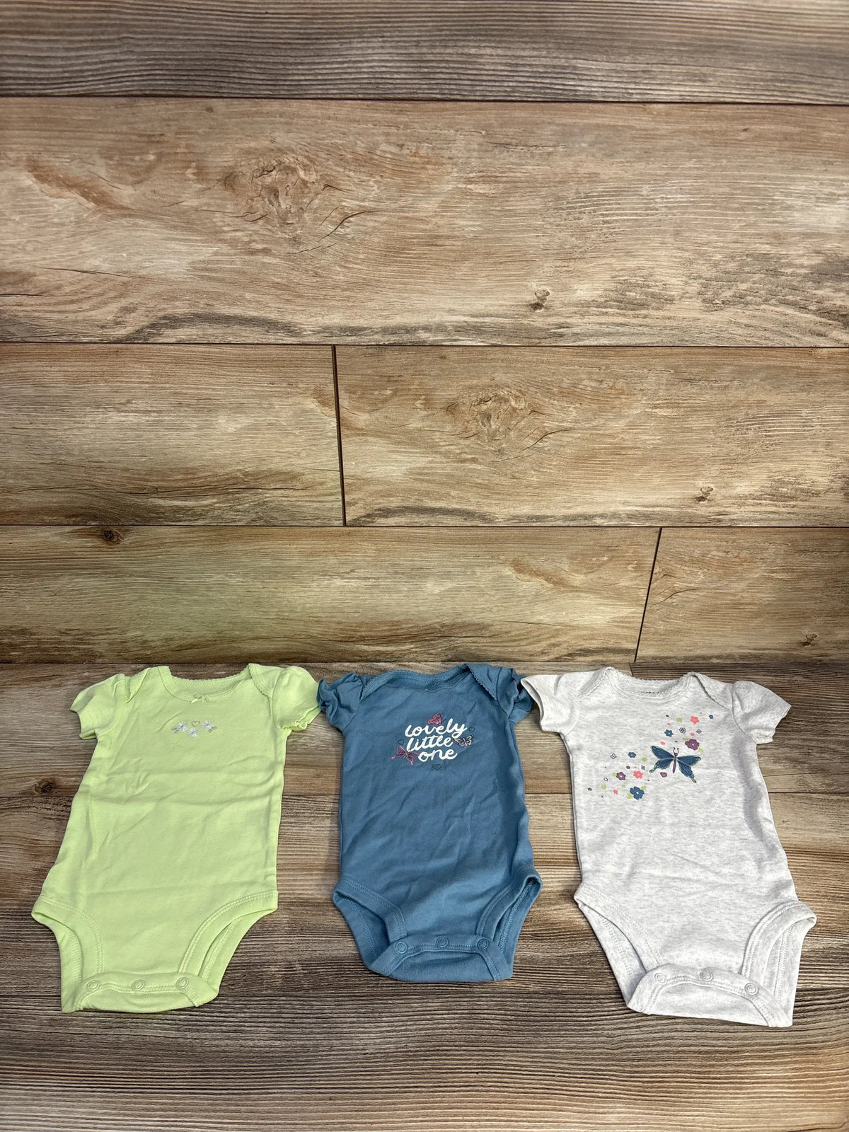 Top view of Carter’s 3-pack bodysuit set showing three short-sleeve bodysuits with floral designs and snap closures on a wood background