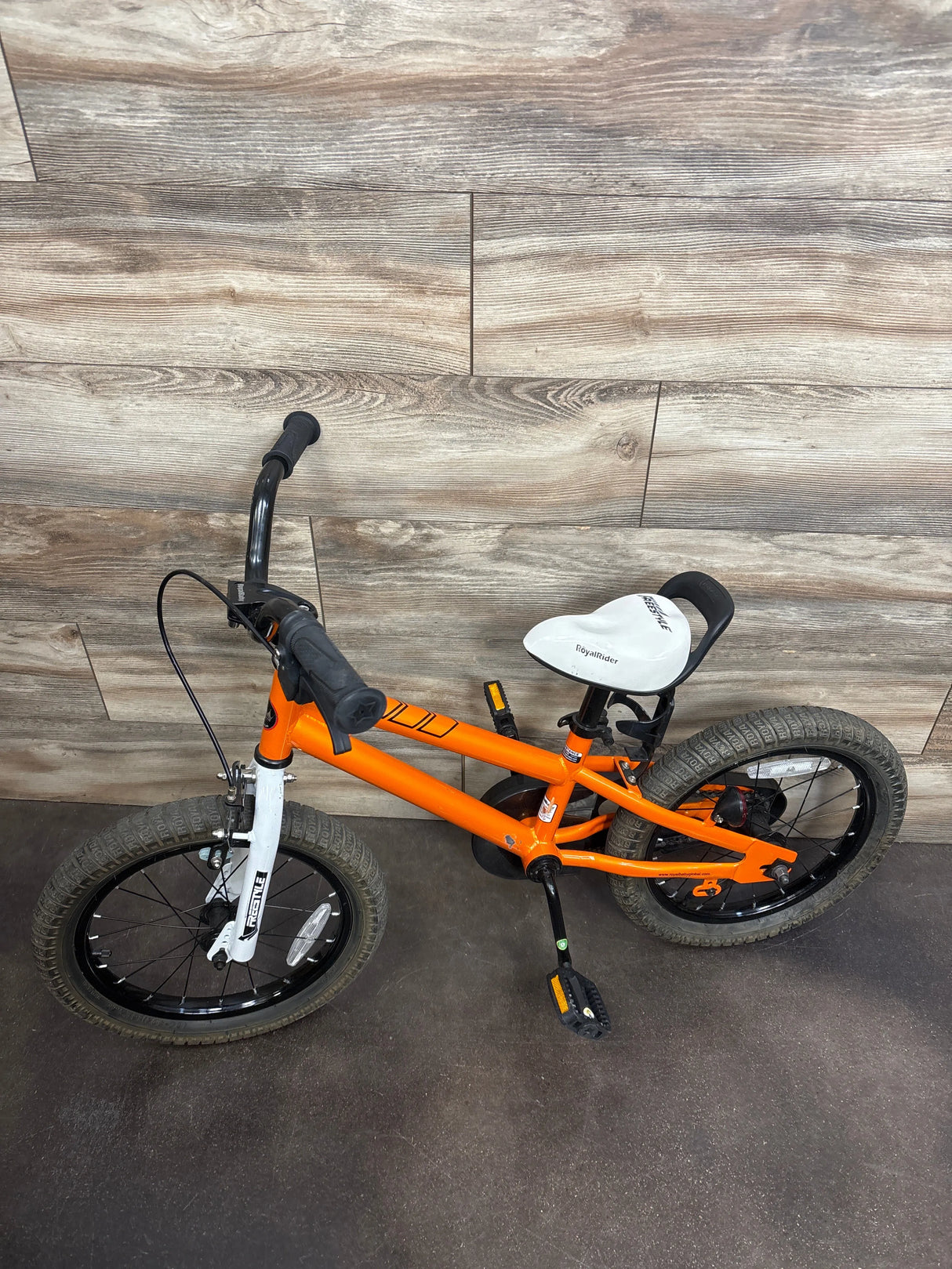 Bright orange kids' bike with training wheels, white seat, and black handlebars on wood floor