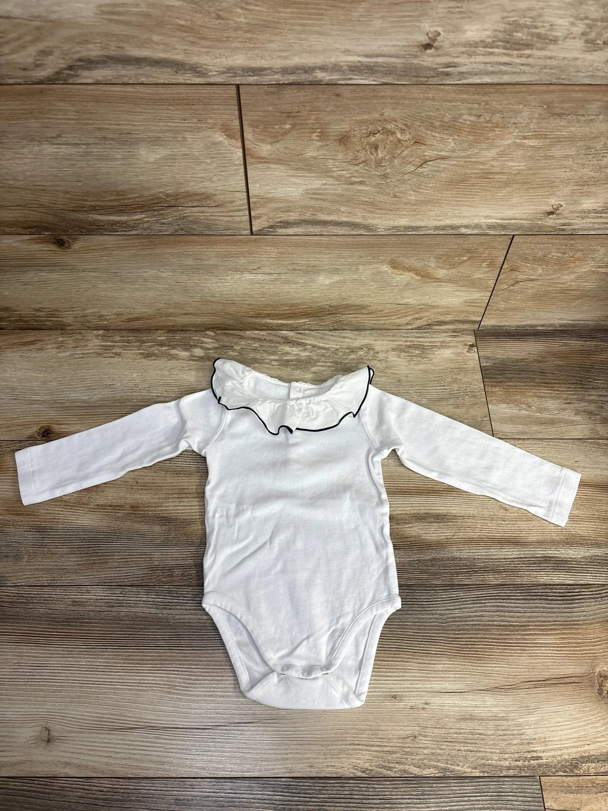 White long-sleeve baby bodysuit with a ruffled collar and black trim on wooden floor