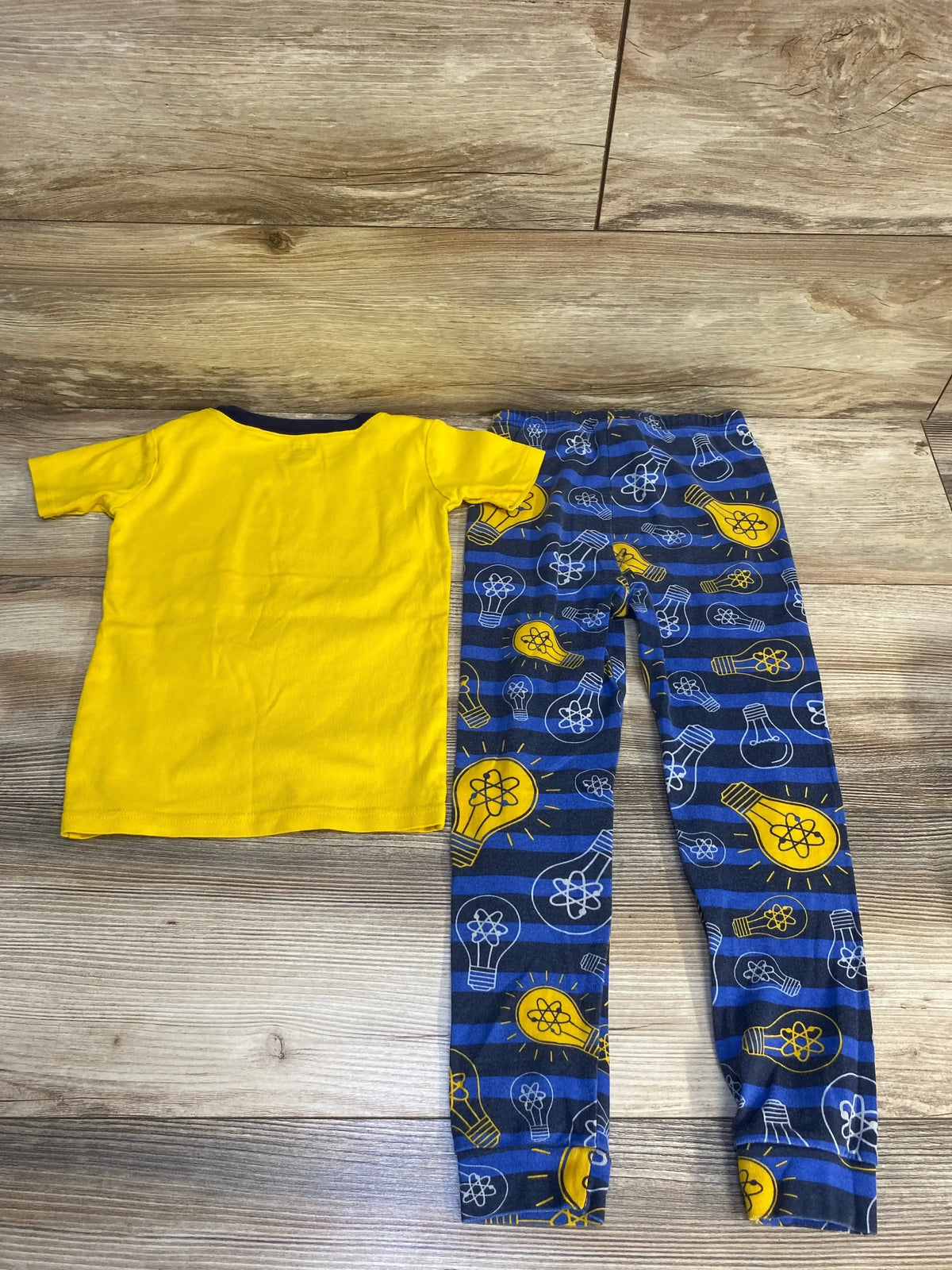 Flat lay of Children’s Place 2pc Genius Like Mom pajama set with yellow tee and blue lightbulb-print leggings on wood floor