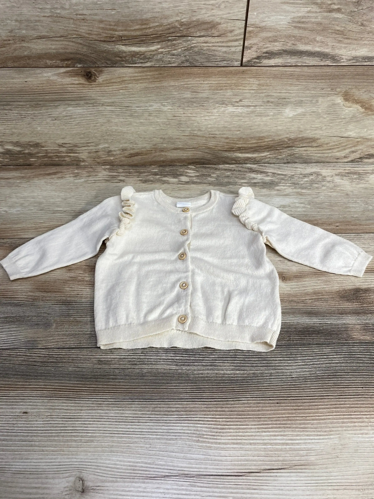 Cream-colored toddler cardigan with wooden buttons and ruffle details on sleeves on wooden floor