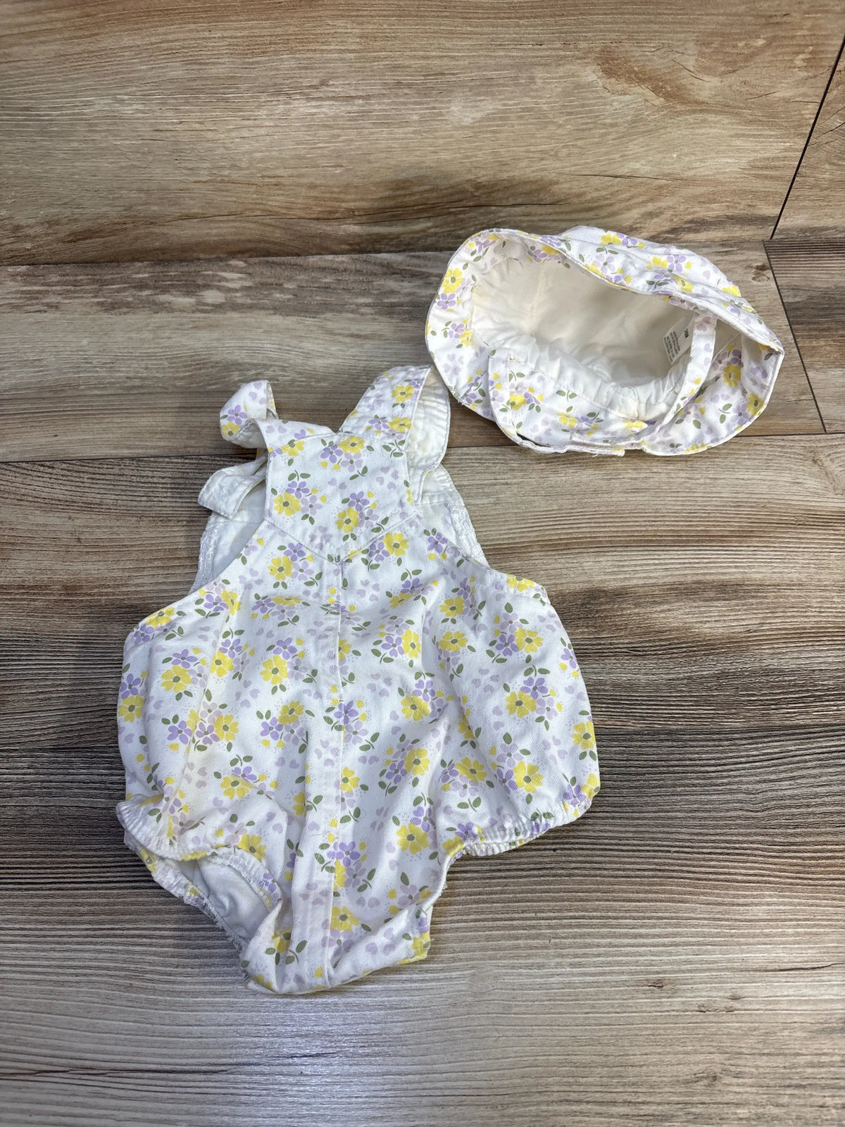 Top view of floral bubble shortalls and matching bucket hat set with yellow and lavender flowers on white, on wood