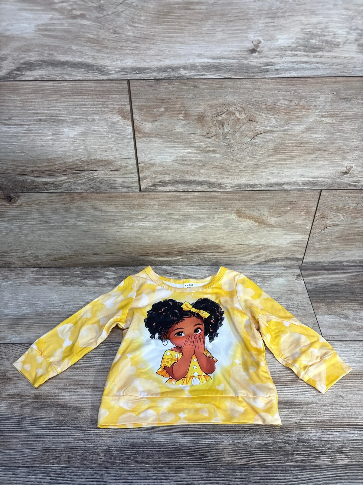 Flat lay of yellow long-sleeve baby shirt (6-9m) showing front cartoon girl with pigtails and bow and white heart print on wood floor