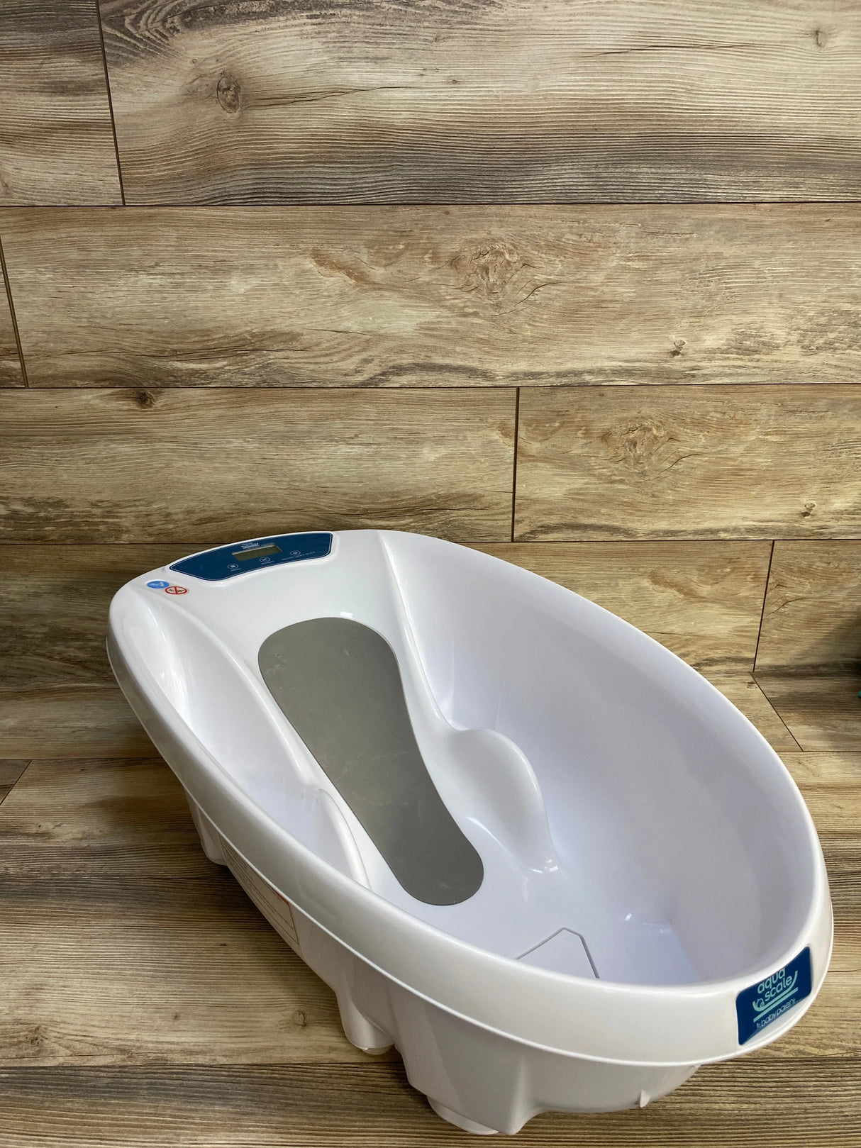 Angled three-quarter view of AquaScale infant tub showcasing digital scale display and gray non-slip seat with wood plank background