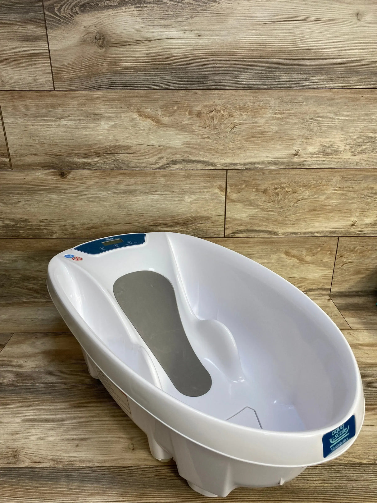 Angled three-quarter view of AquaScale infant tub showcasing digital scale display and gray non-slip seat with wood plank background