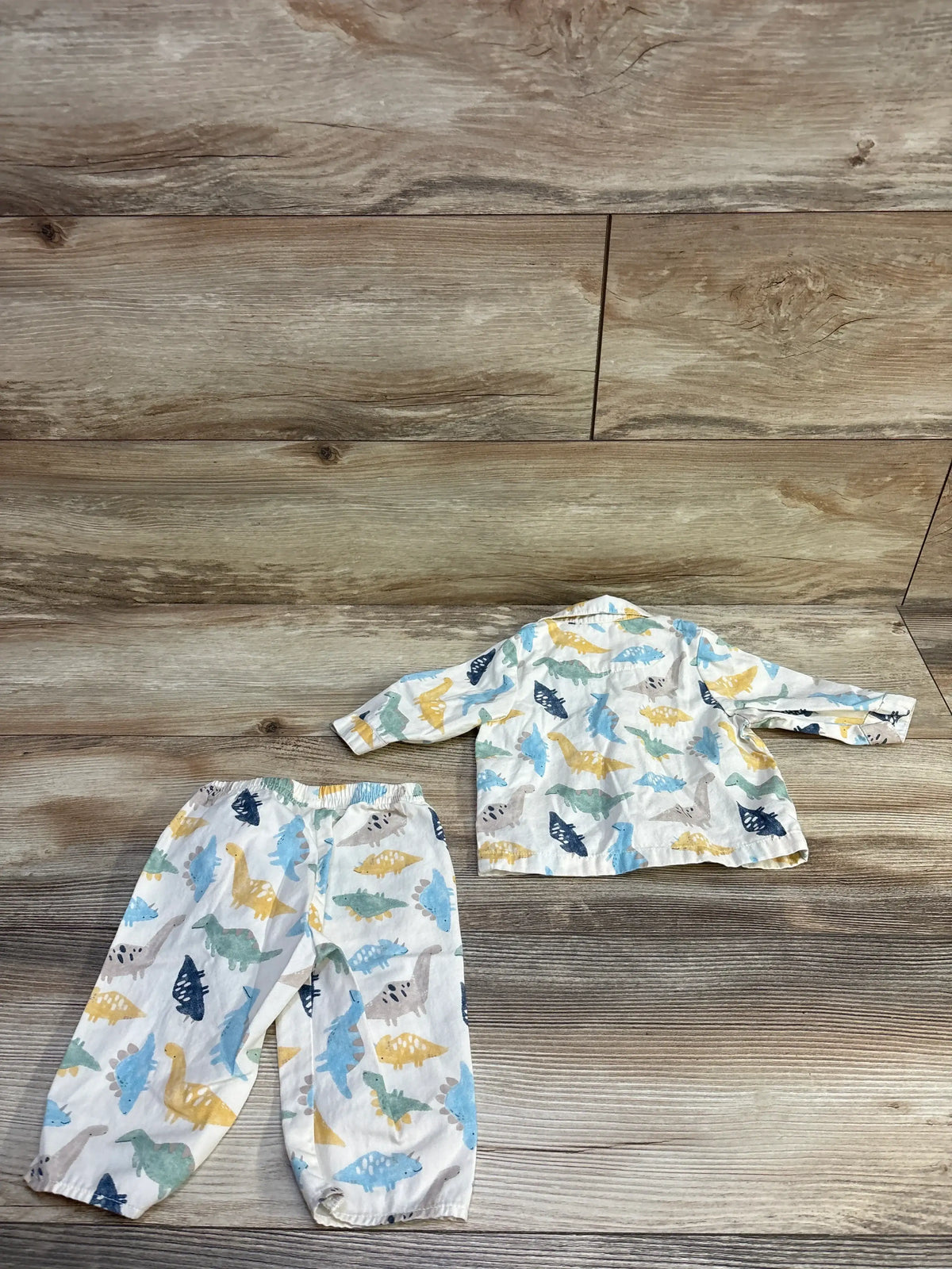 Flat lay of ivory 2-piece baby button-up shirt and elastic-waist pants showing multicolored dinosaur print on wood background