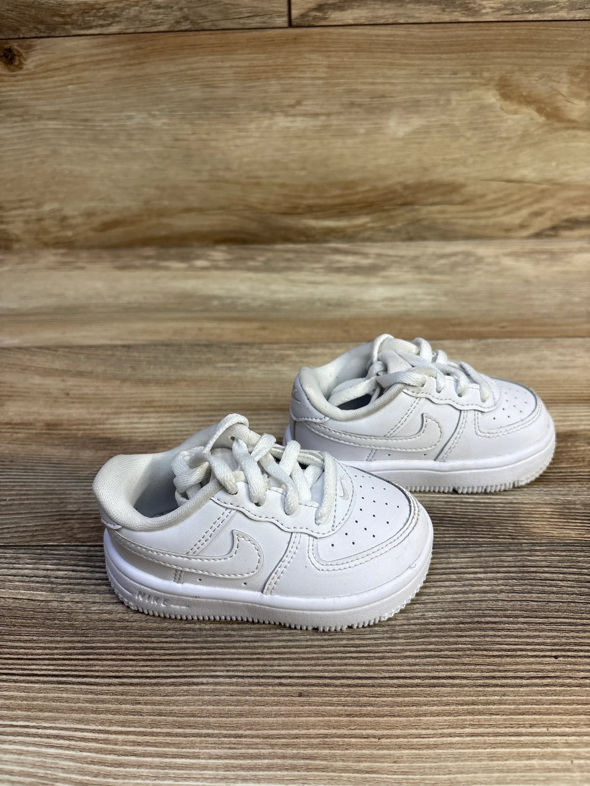 Three quarter view of toddler white Nike Air Force 1 Low sneakers size 5C showcasing stitched Nike Swoosh and perforated toe on wood background