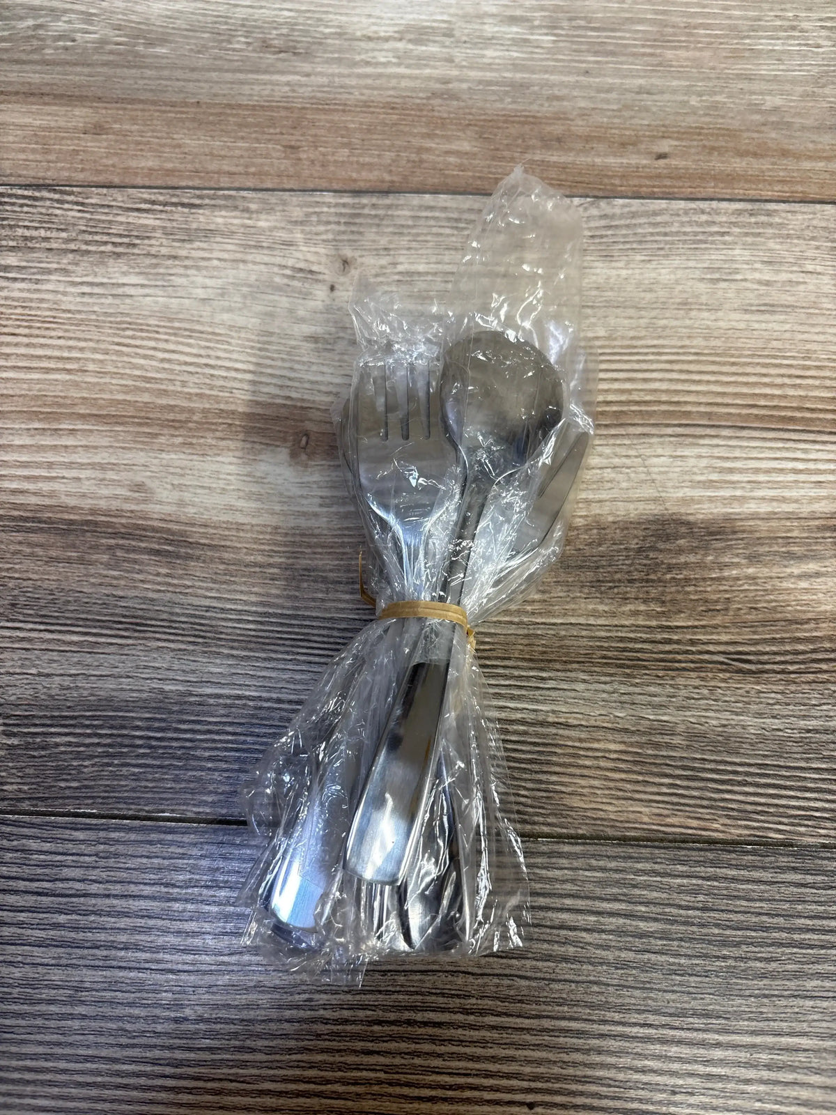 Top-down view of a 10-piece stainless steel toddler utensil set wrapped in clear plastic and secured with a rubber band on a woodgrain background