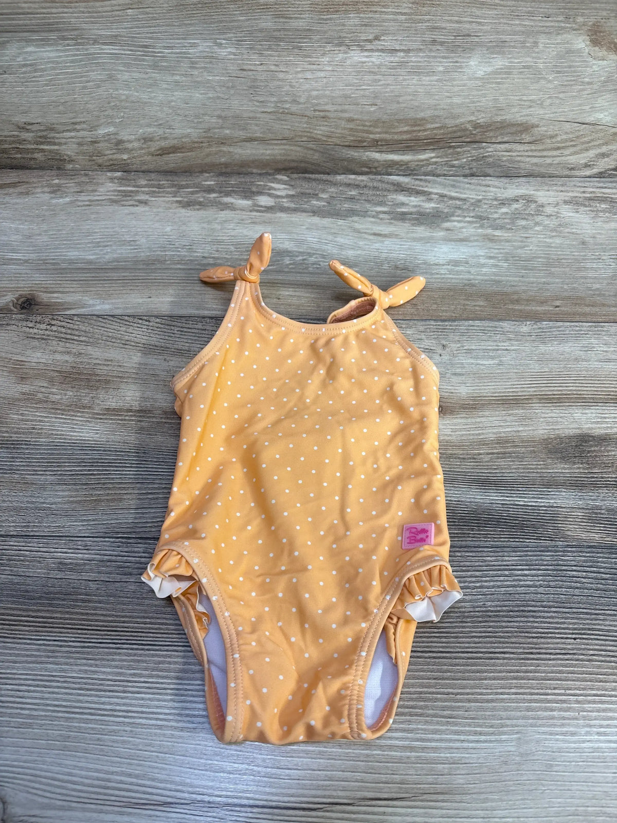 Flat lay of Ruffle Butts baby one-piece swimsuit, melon orange with white polka dots, tied shoulder straps and ruffled legs on wood