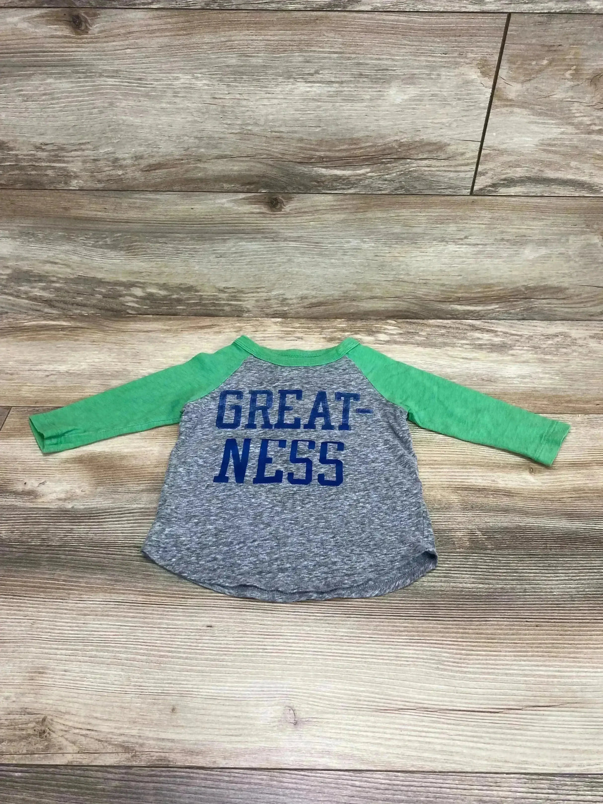 Flat lay of Crewcuts Like Lightning Raglan Shirt Green and Grey sz 2T showcasing GREATNESS text graphic with green sleeves on wood plank floor