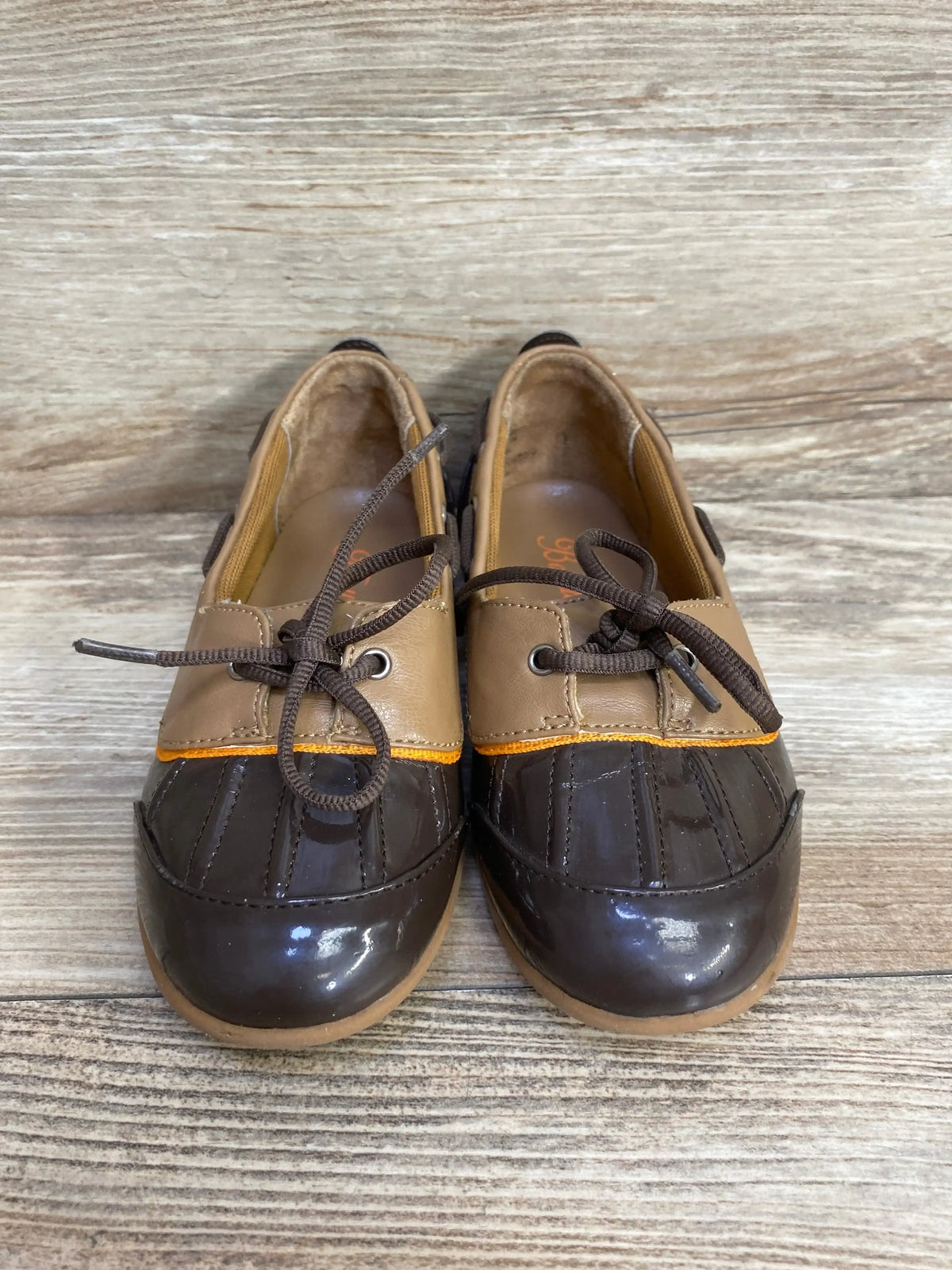 Front view of Balleto brown boat shoes showing dark patent toe, tan upper with orange trim and tied laces on wood background