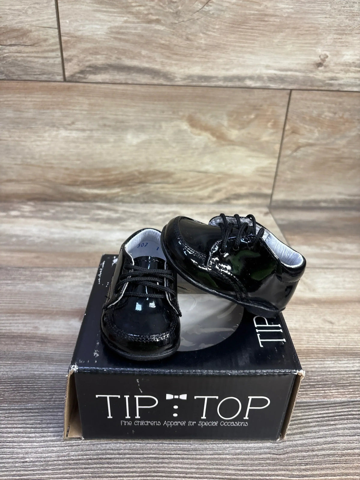 Three quarter view of Tip Top black patent leather baby sneakers showing lace up detail and glossy finish on branded box
