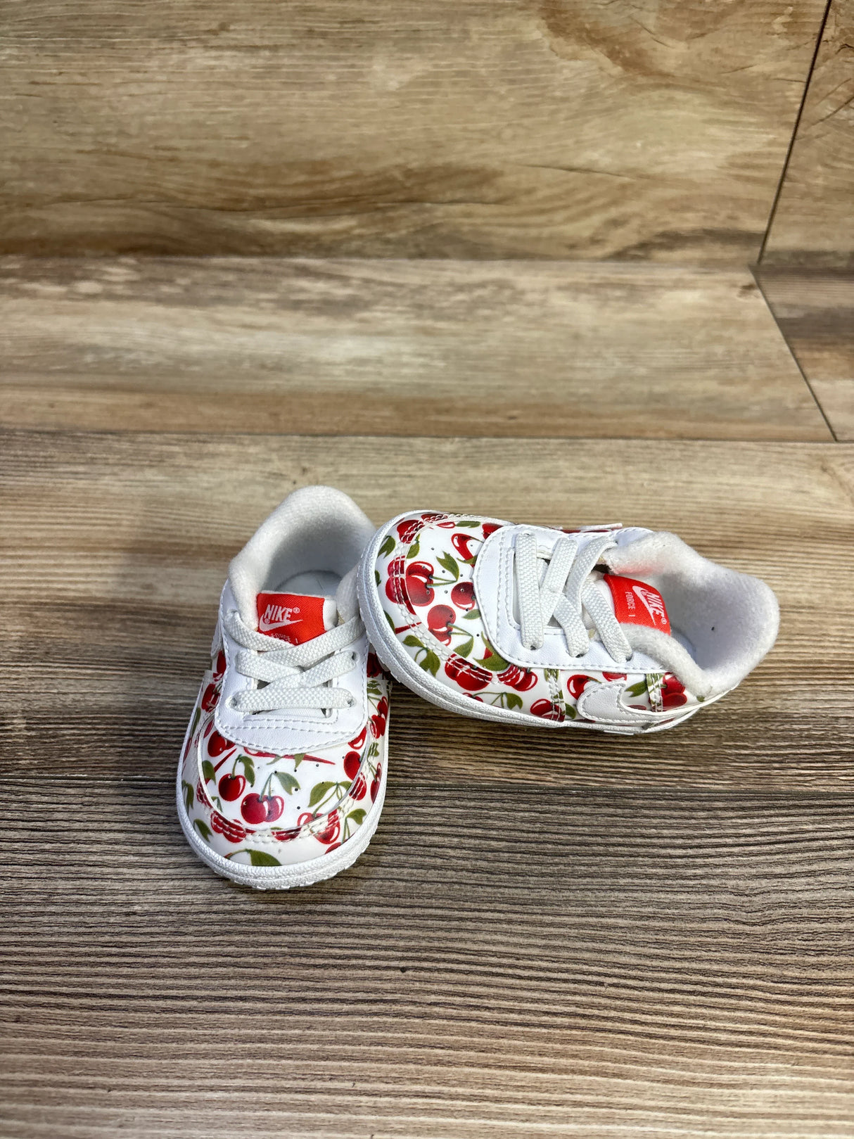Front view of Nike Air Force crib shoes showcasing red cherry print on white leather and lace-up front on wood surface