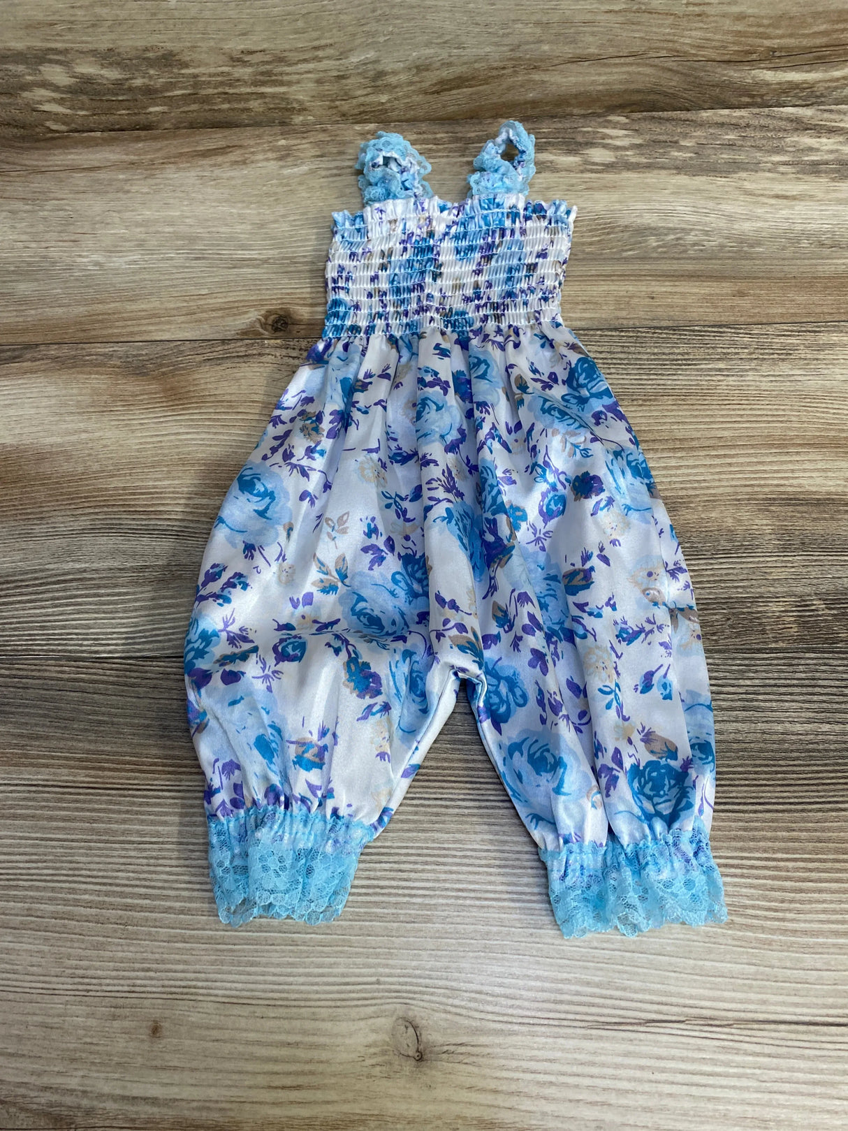 Flat-lay of white floral smocked baby romper (3M) showing smocked bodice, blue floral print and lace-trimmed ankles on wood background