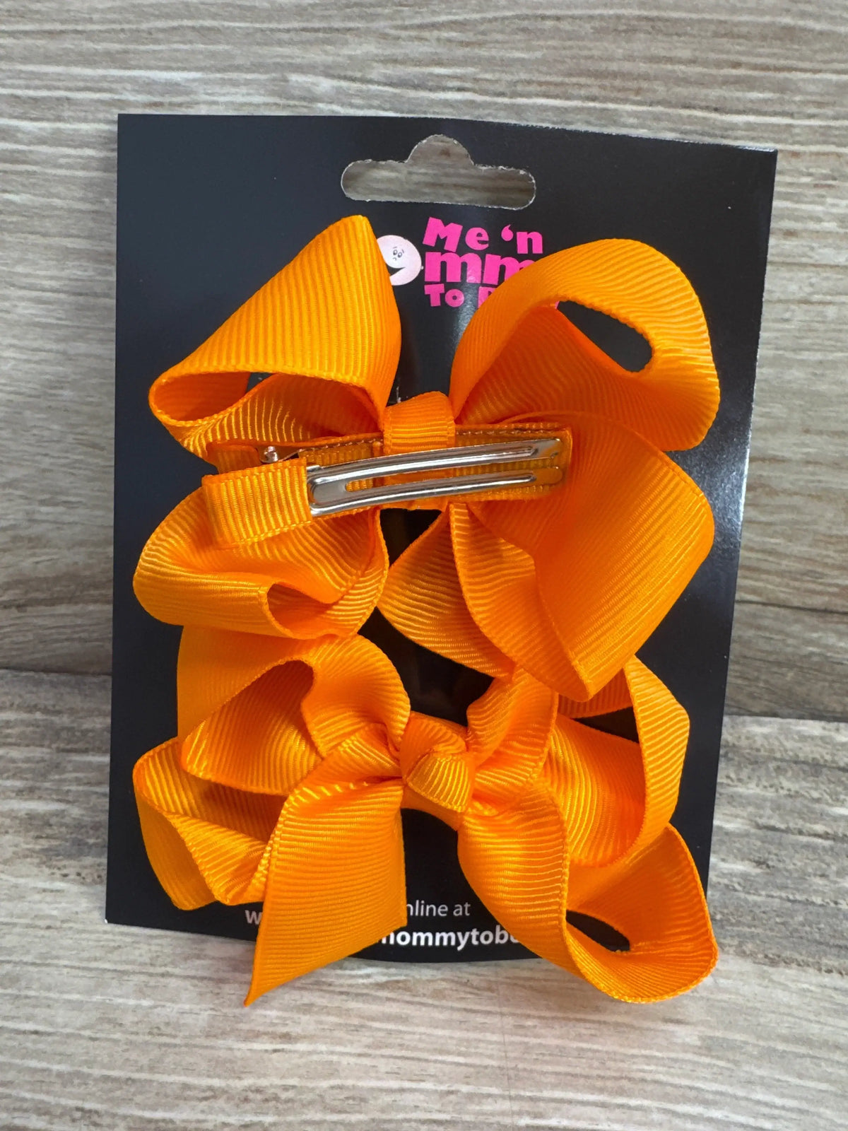 Two medium orange ribbon hair bows on metal alligator clips attached to black display card on woodgrain background