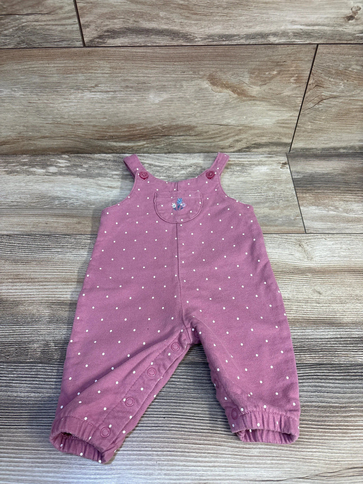 Front view of pink polka-dot baby overalls with embroidered chest pocket, snap inseam closures and button straps on wood floor