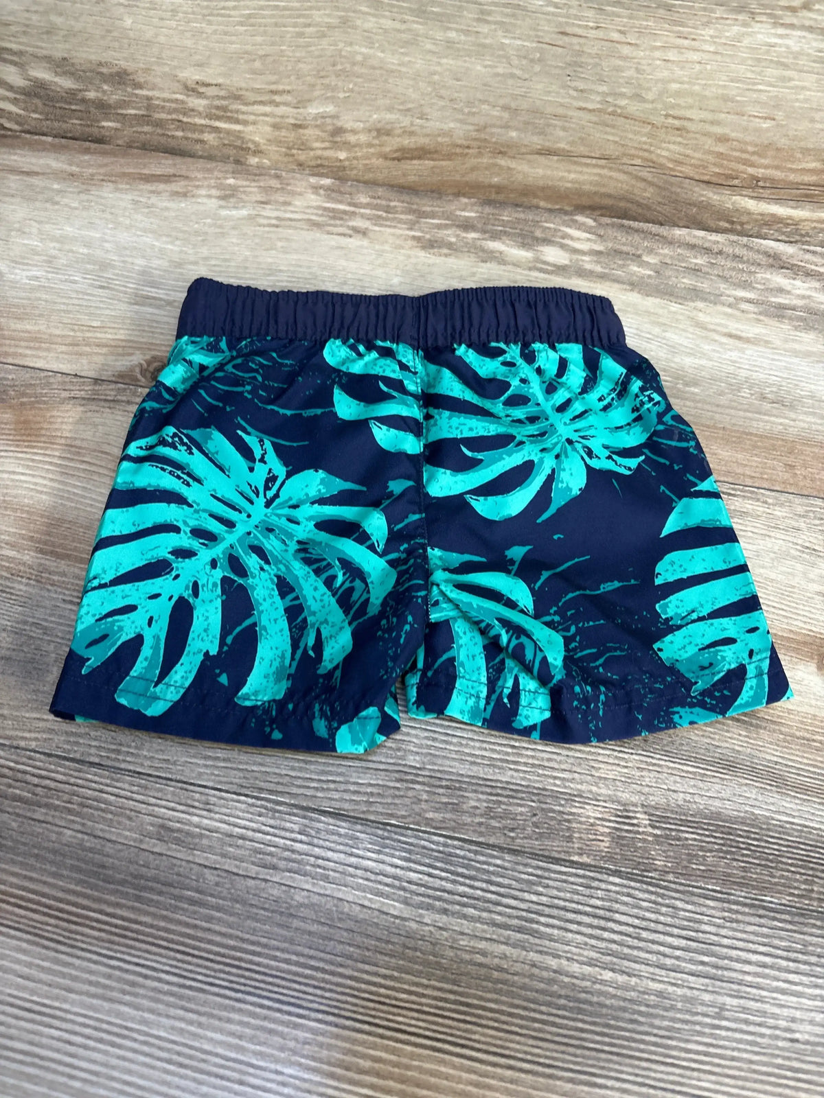 Flat lay of Cat & Jack swim trunks (12 months) showcasing teal tropical leaf print and elastic waistband on wood floor background