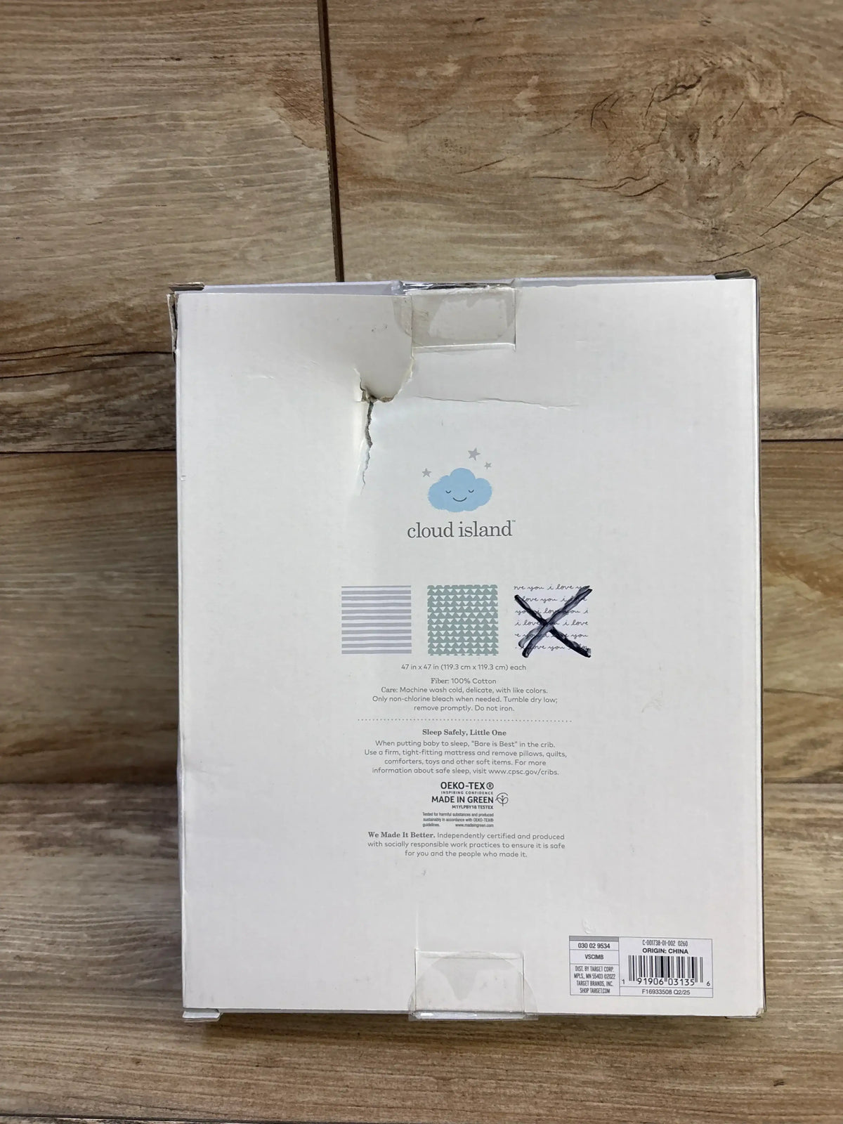 Back of Cloud Island 2-pack muslin swaddle box showing brand logo, three pattern swatches (one crossed out), torn top flap, on wood surface