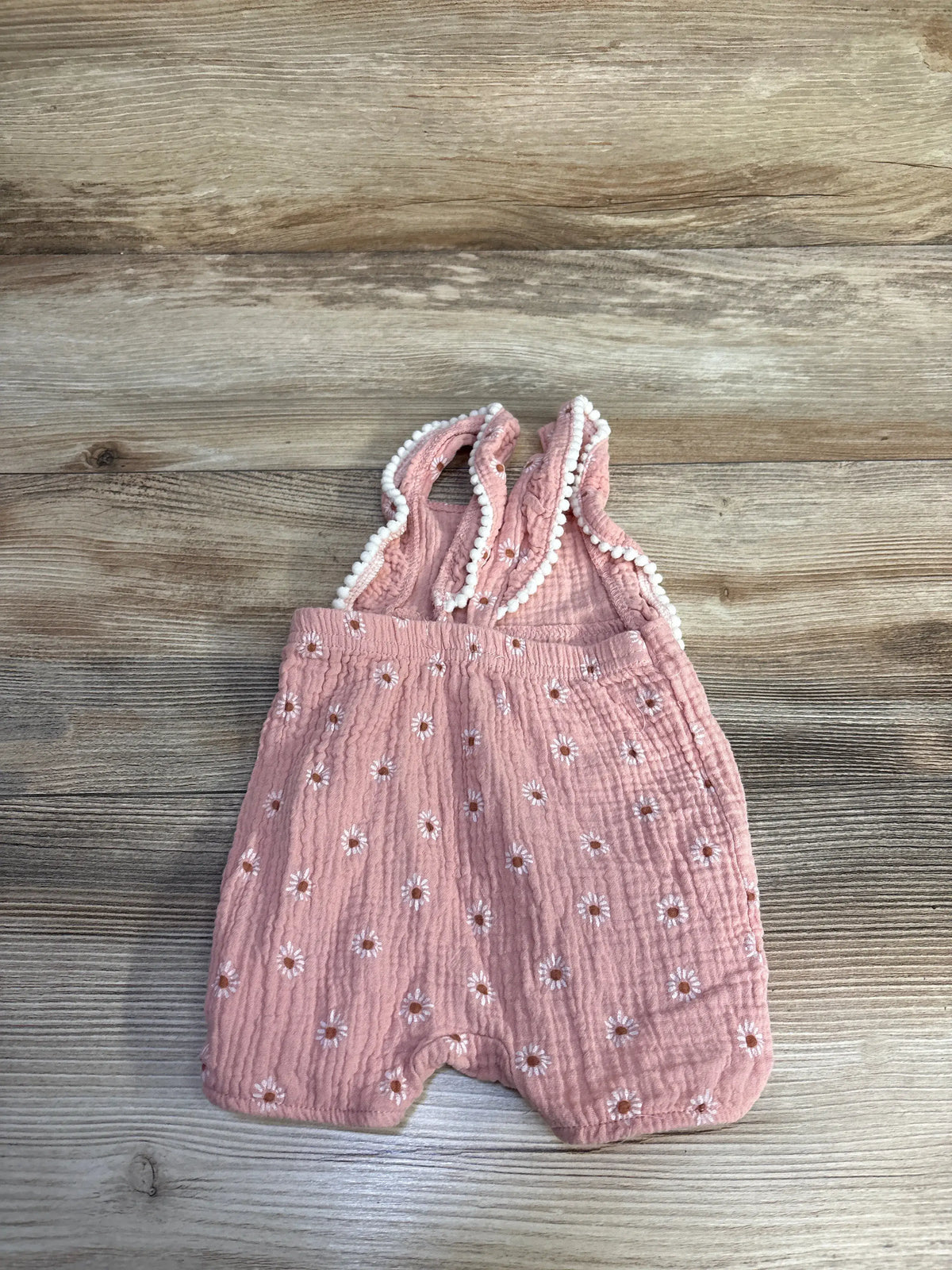 Front view of Emily & Oliver pink floral gauze romper showcasing floral print and white pom-pom trim with wood background