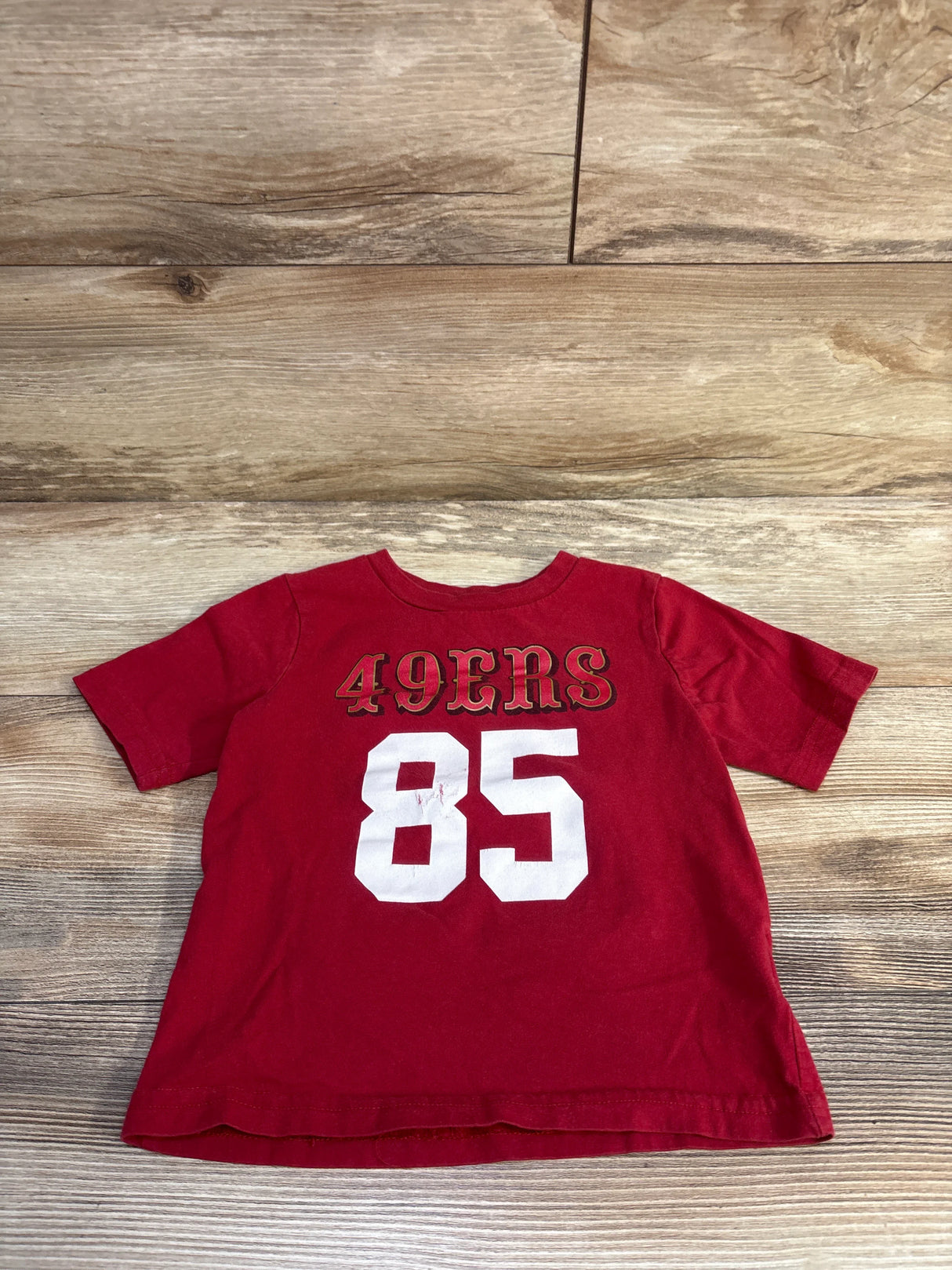 Flat-lay of red toddler 49ers T-shirt showing white 85 and gold-outlined 49ERS text on wood background