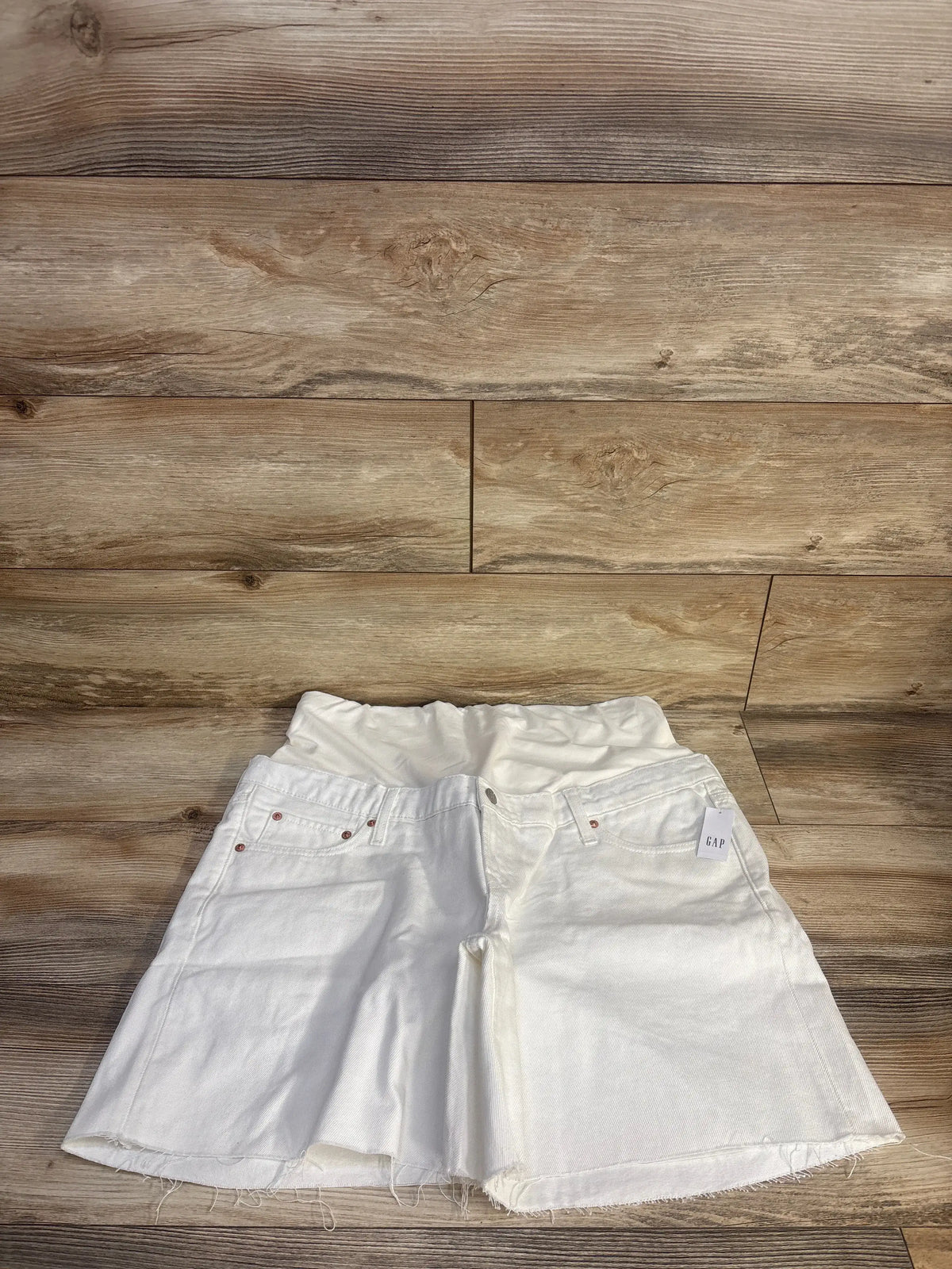 Front view of white Gap maternity Bermuda shorts showing full panel, five-pocket styling, and frayed hems on wood flooring