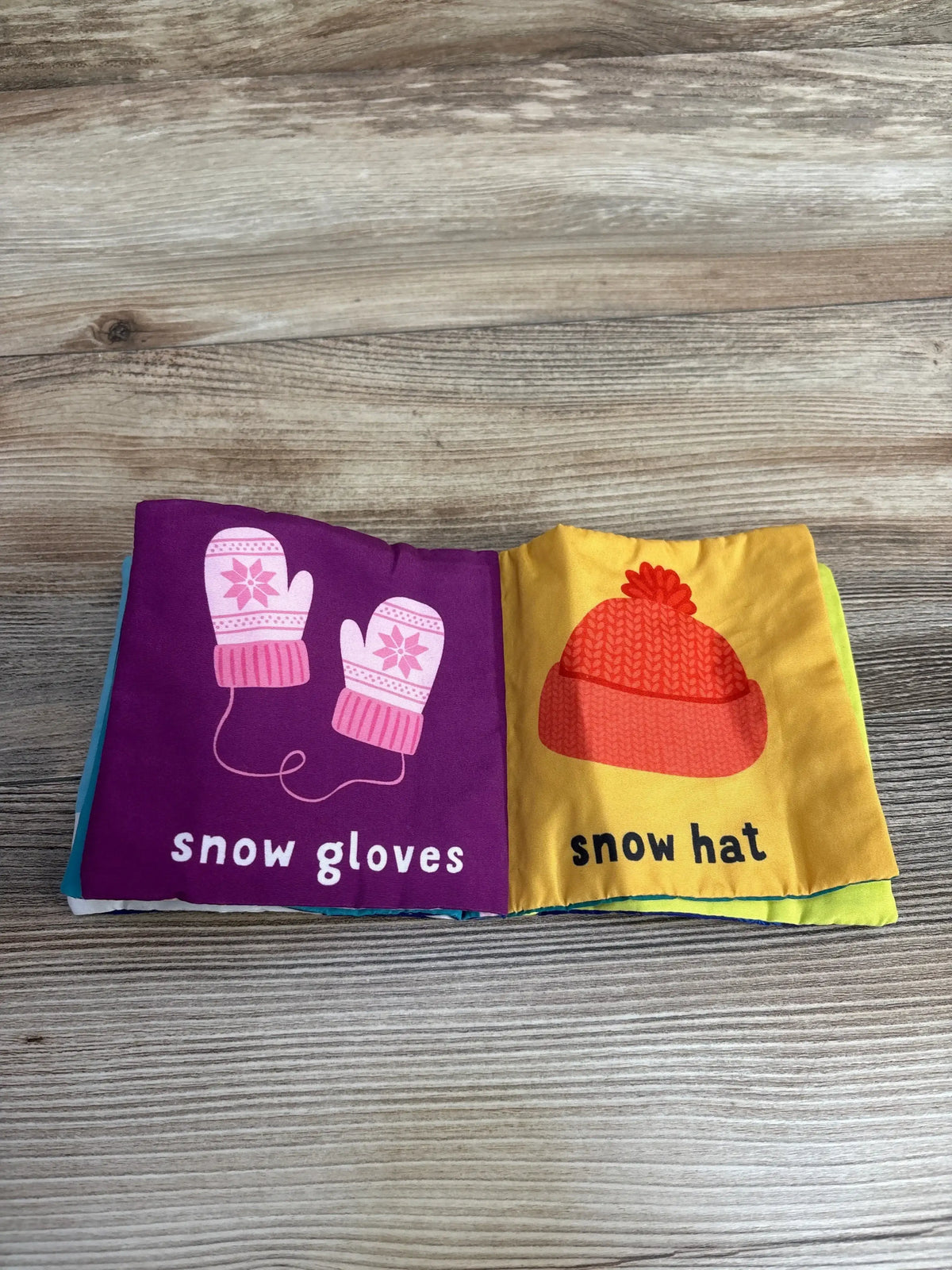 Soft baby cloth book open to snow gloves and snow hat illustrations on wood surface