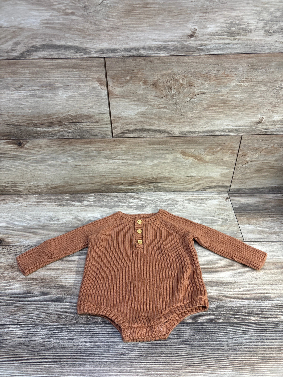 Front view of Little Planet brown knit henley bodysuit with long sleeves, ribbing, three buttons, snaps, and wood backdrop