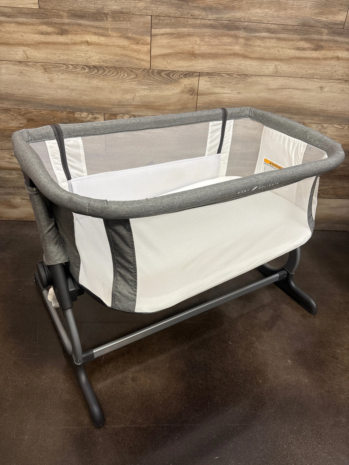Three-quarter view of Baby Delight Beside Me Dreamer bedside bassinet in grey showcasing mesh sides with a wood wall background