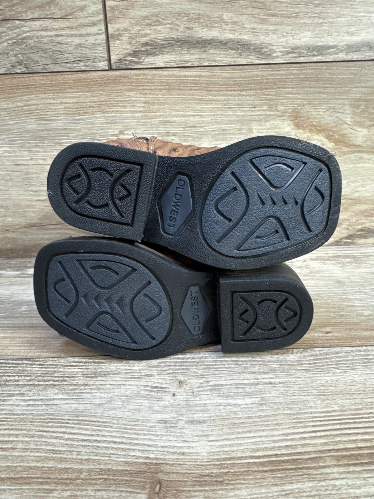 Bottom view of Old West ostrich-print boots showing soles with tread pattern and ‘OLD WEST’ logo on wood floor