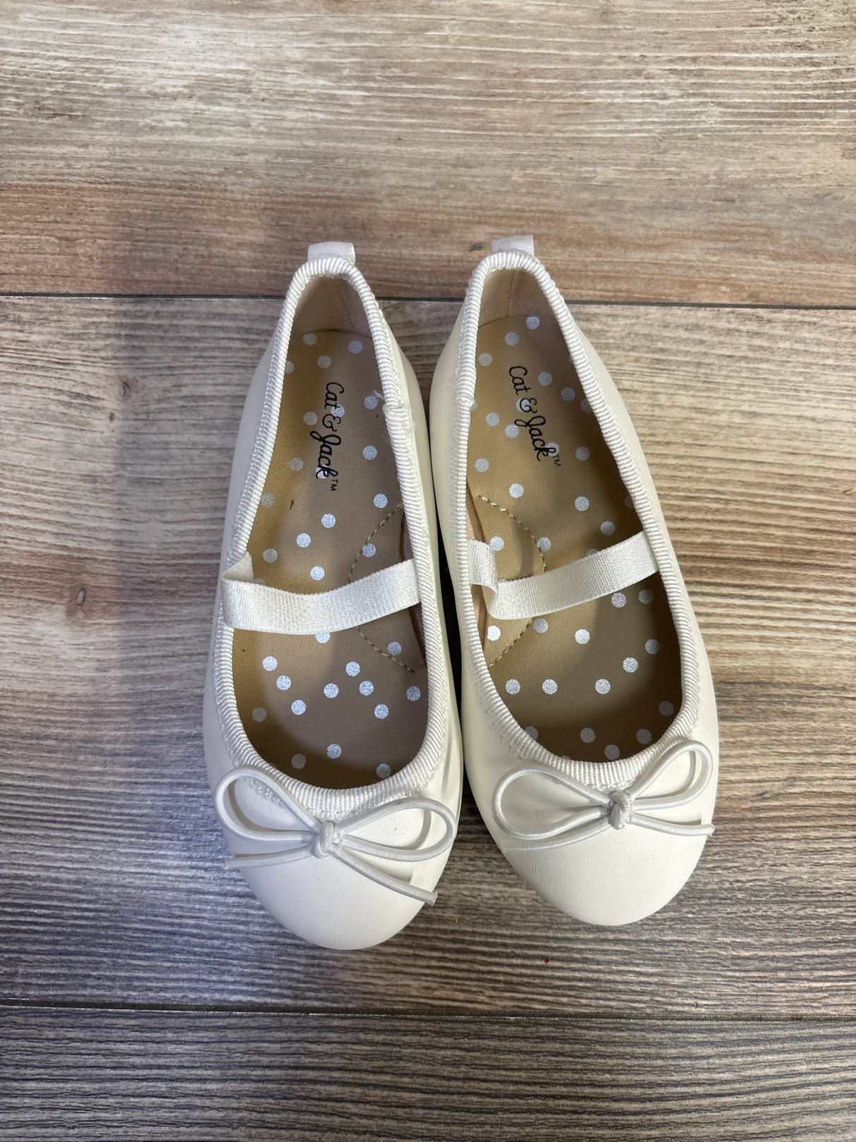 Top view of white Cat & Jack Nora slip-on ballet flats with bow detail, elastic strap, and polka-dot insoles on wood floor