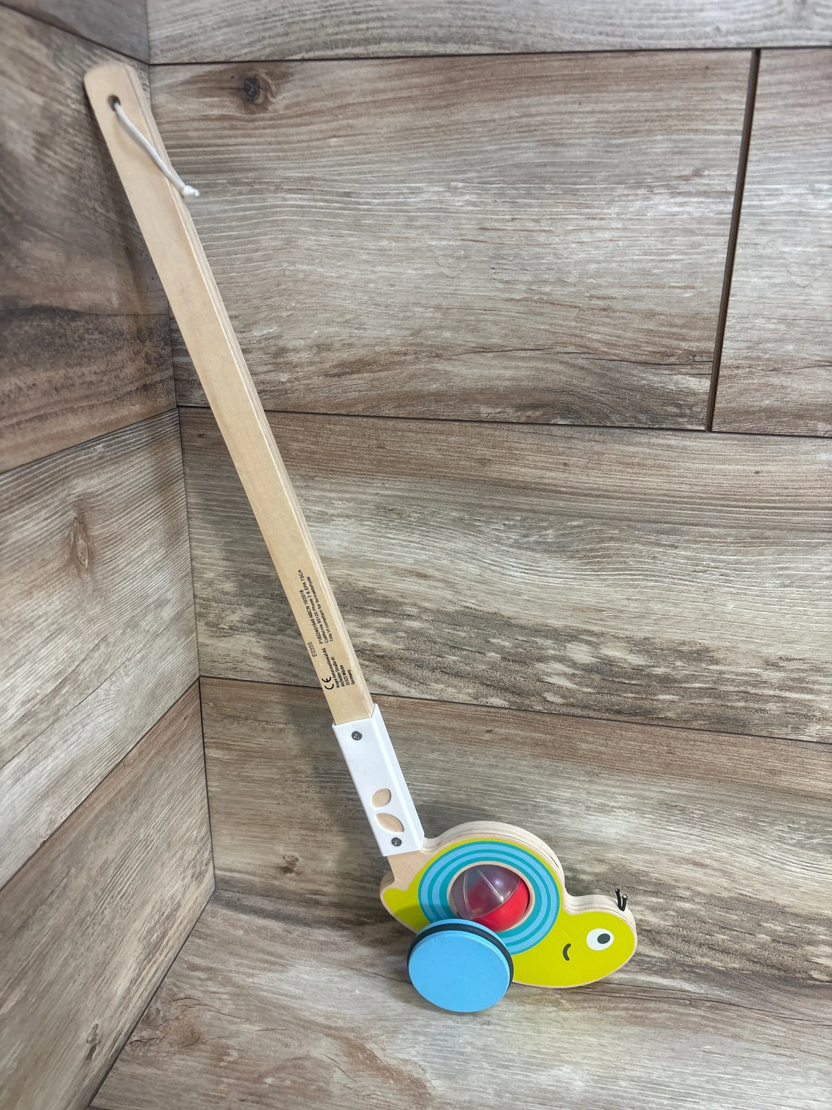 Angled view of Hape Snail Push Pal walking toy showing long wooden handle, blue wheels, and colorful spinning ball on wood floor