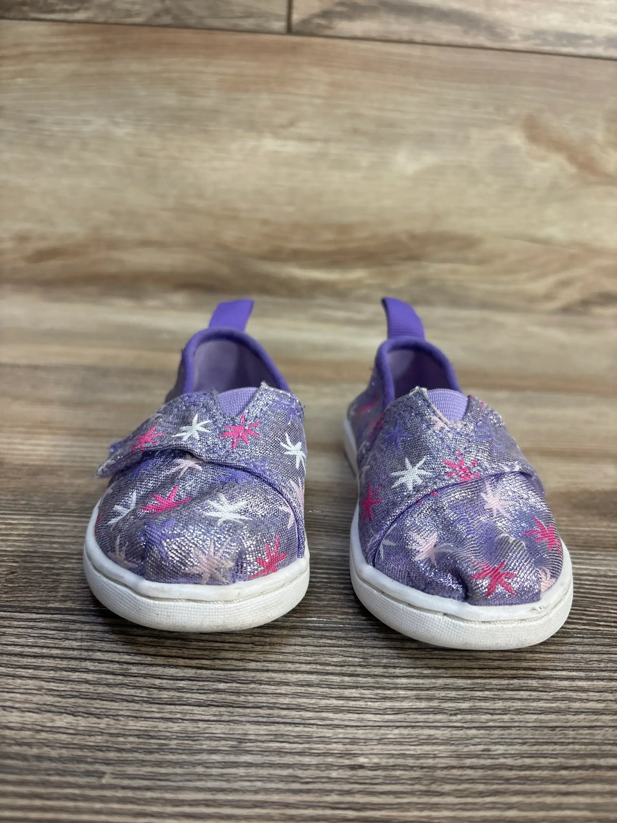 Purple kids' slip-on sneakers with starfish print, glitter fabric, and white soles on wood floor