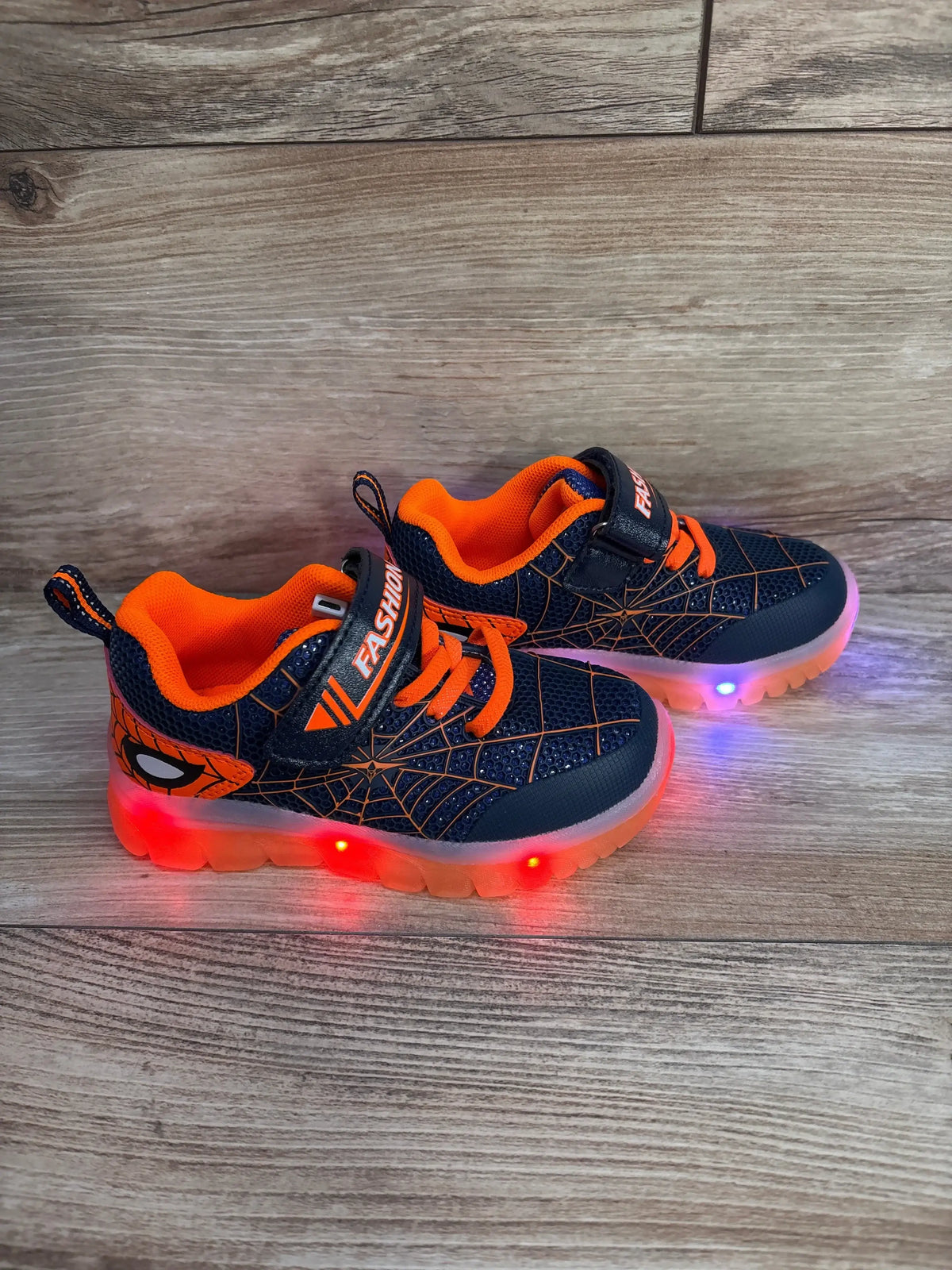 Side view of Jian Dian navy-orange toddler light-up sneakers with spiderweb pattern, Velcro strap and glowing sole on wood