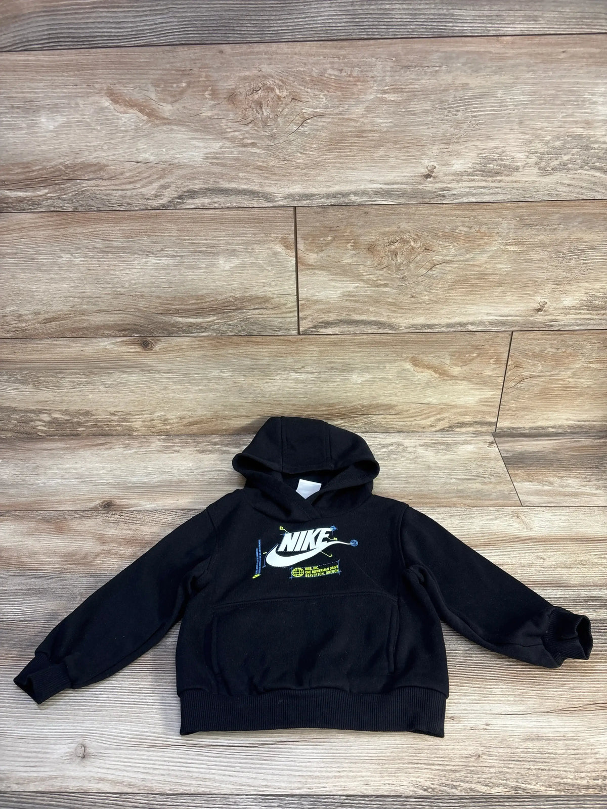 Front view of black Nike toddler hoodie 24m with white logo, kangaroo pocket and ribbed cuffs on wood floor