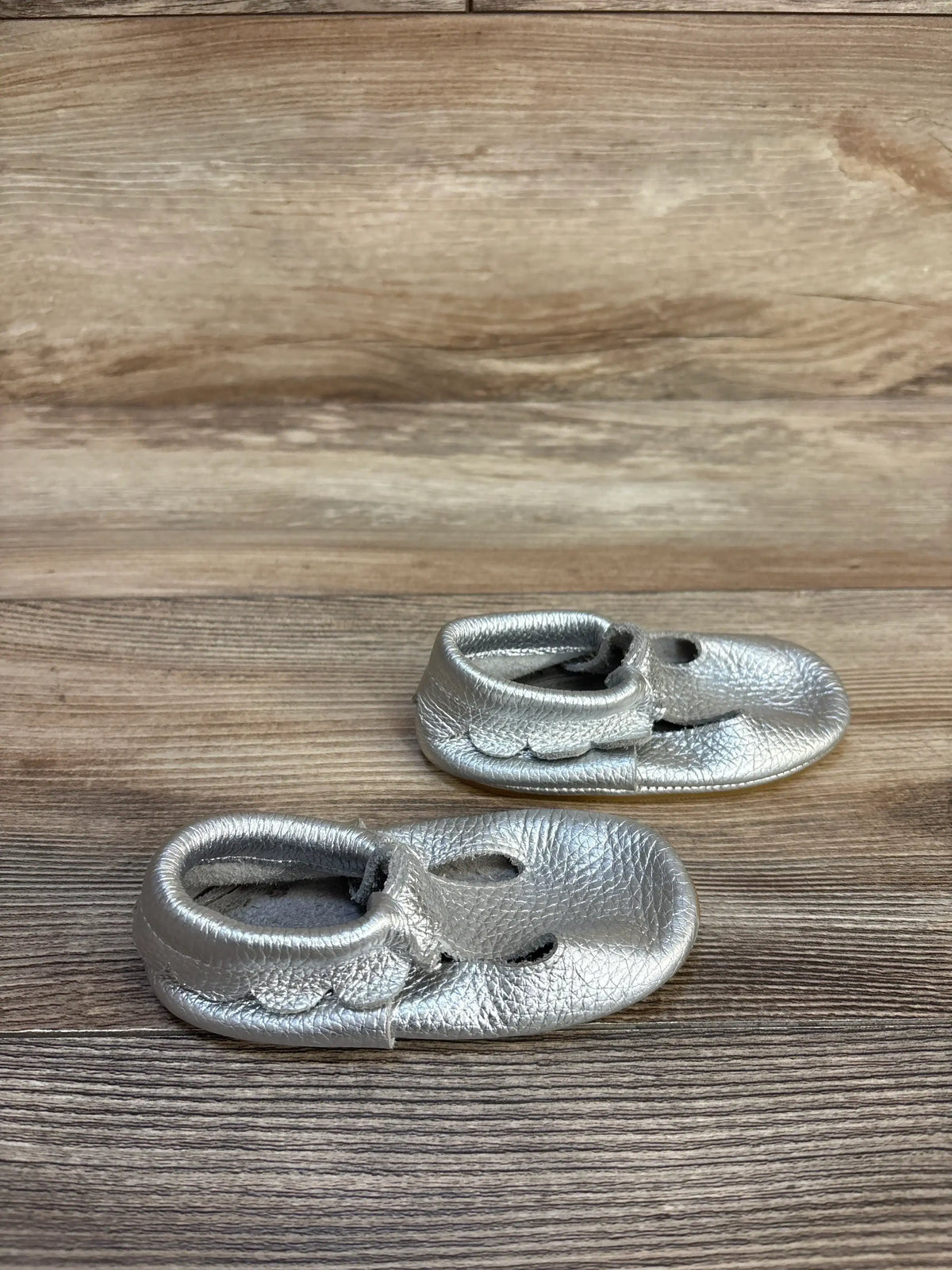 Silver baby moccasins with scalloped edges on wooden background, kids footwear